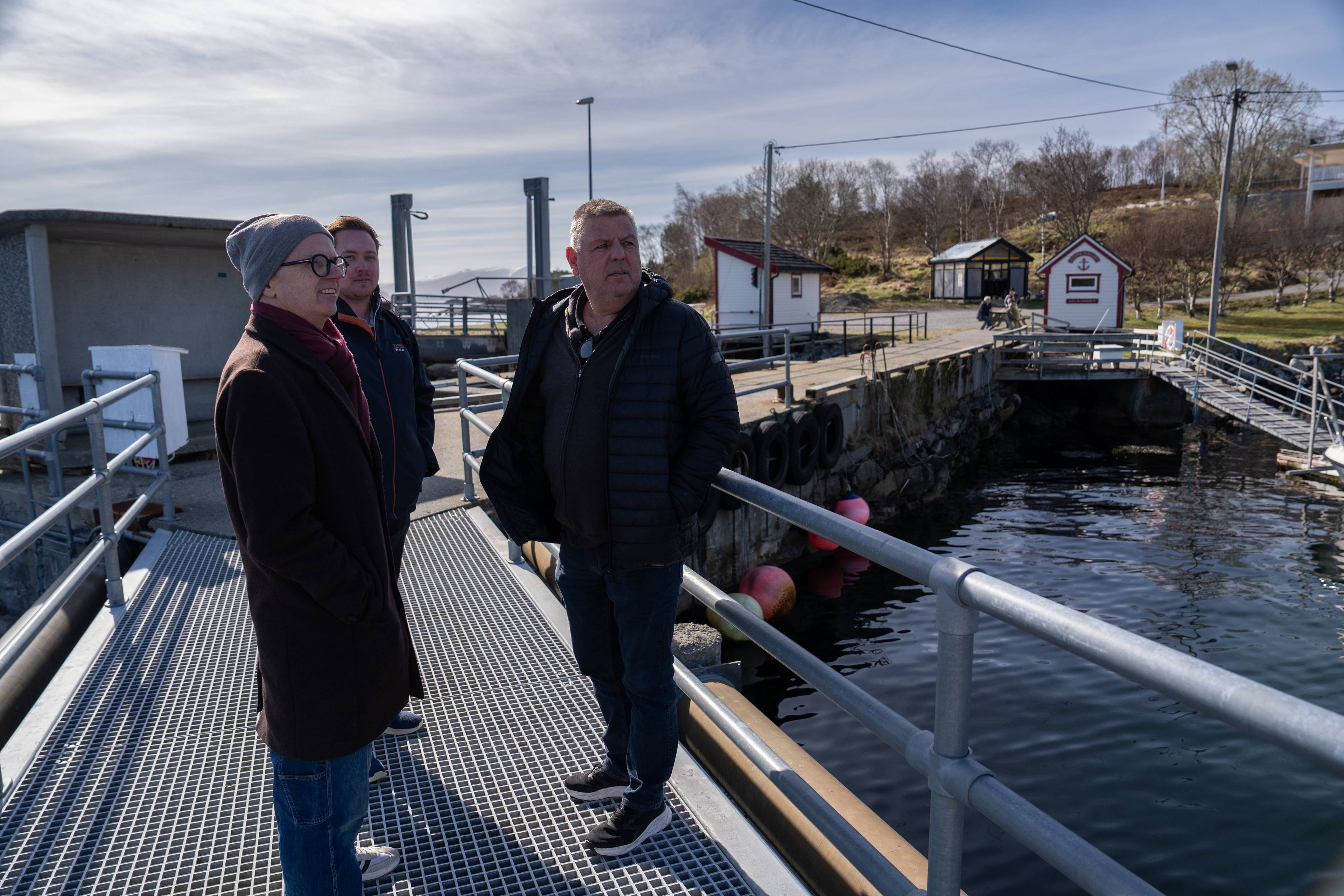 Tre menn står på en brygge ved siden av et havnebasseng. En av mennene er kledd i en brun frakk og et grått skjerf. Den andre mannen, som står i midten, har på seg en svart boblejakke. Den tredje mannen, som står til høyre, har også en svart jakke på seg. Bryggen er laget av metallgitter og har rekkverk. Det er noen hus og trær i bakgrunnen. (Bildebeskrivelsen er laget av en KI-tjeneste)