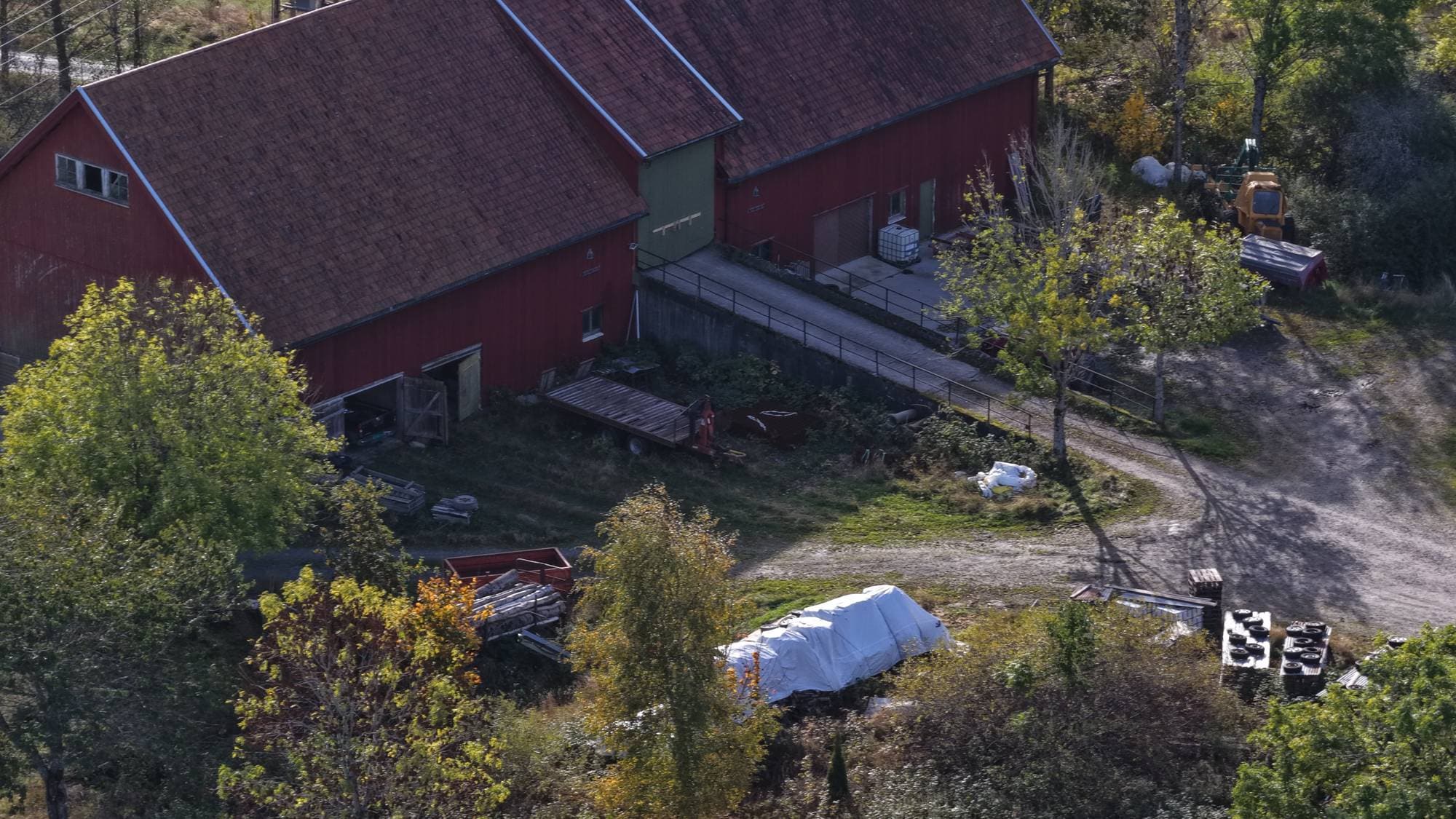 Luftfoto av en rød låve med gårdstun og utstyr på et område med trær og vegetasjon. (Bildebeskrivelsen er laget av en KI-tjeneste)