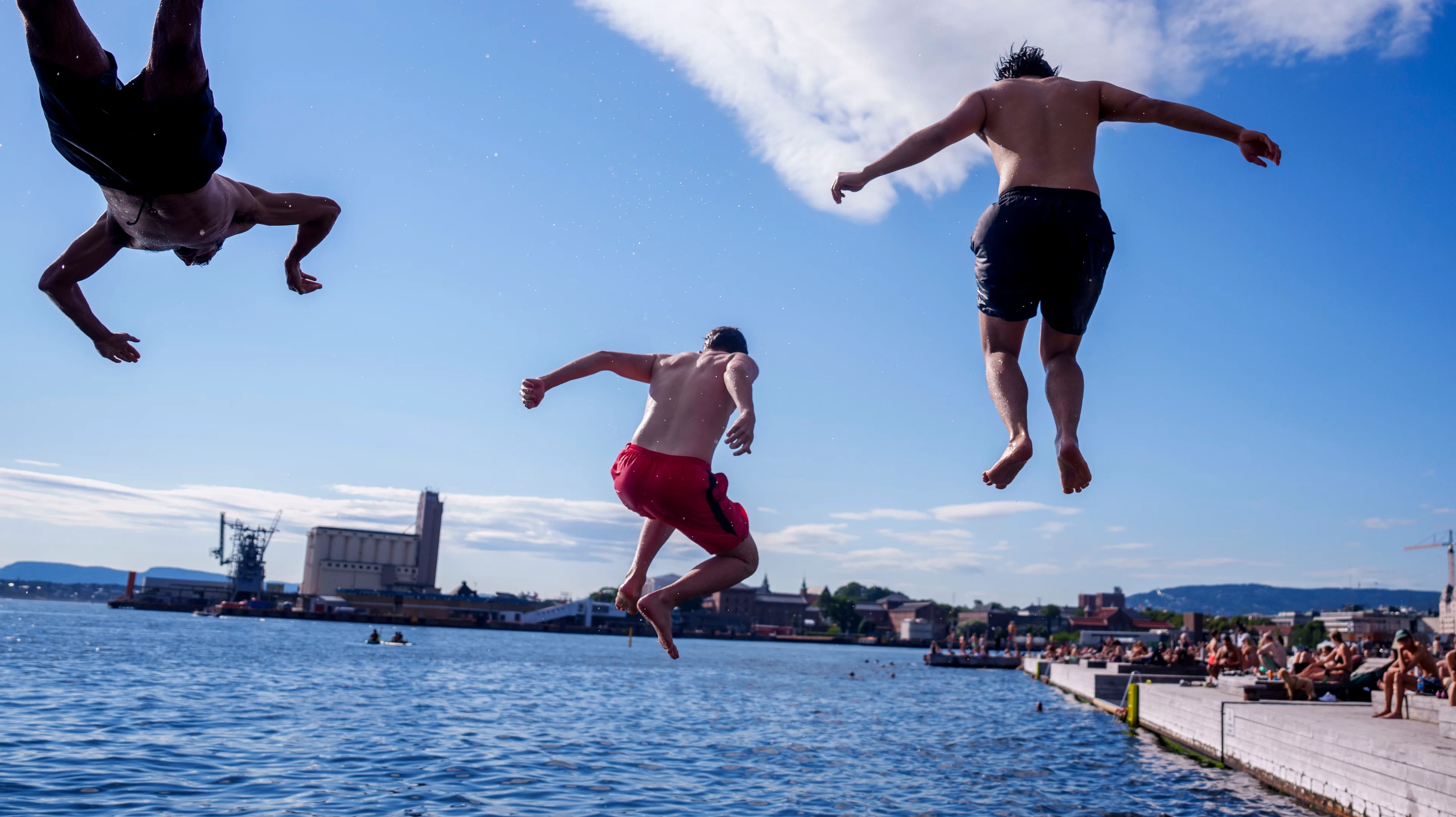 Tre personer hopper ut i vannet fra en badeplass i Oslo med industribygg i bakgrunnen.