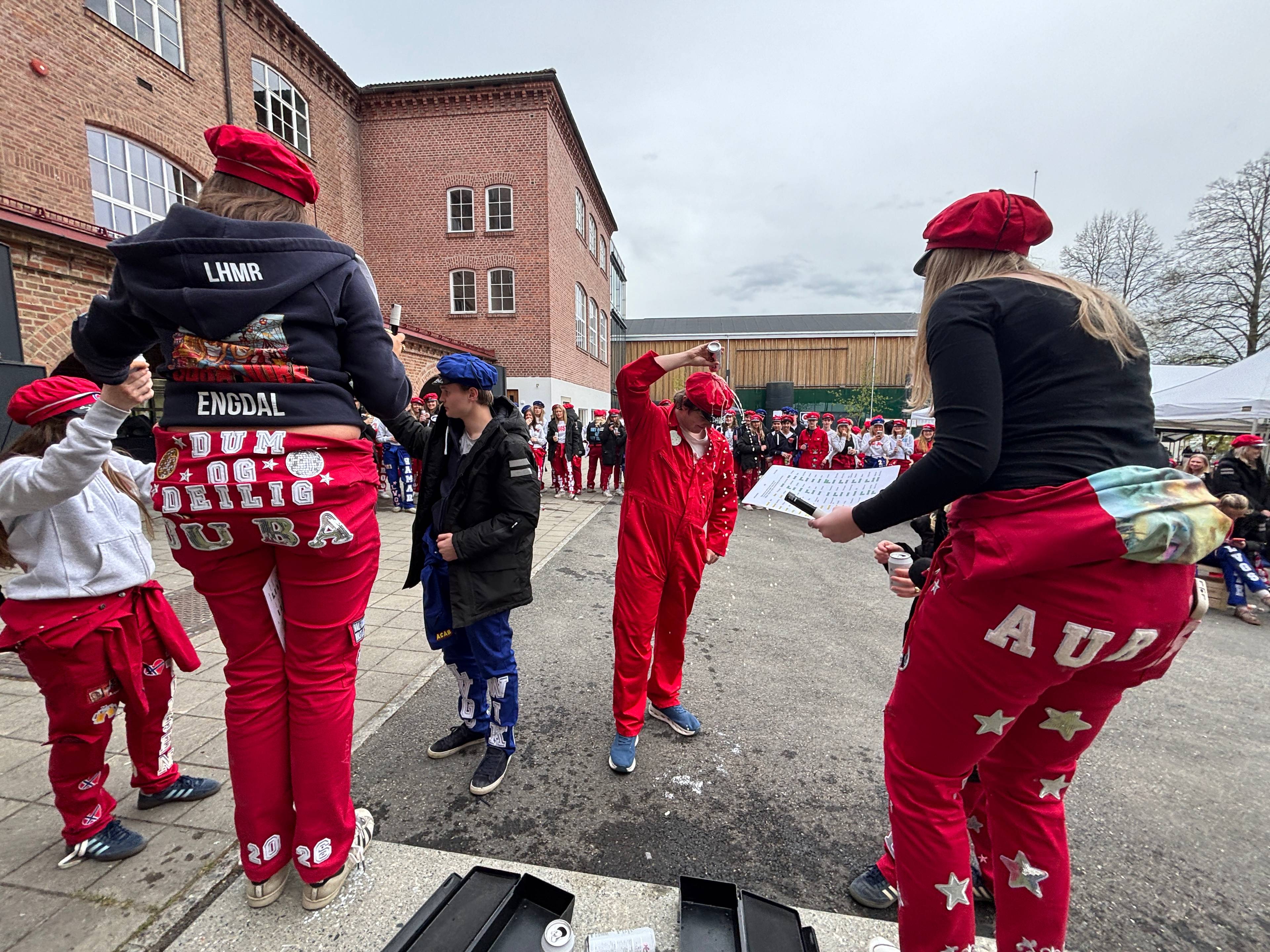 Flere unge mennesker kledd i røde og blå russedrakter står ute. Flere har på seg røde luer og røde kjeledresser.
