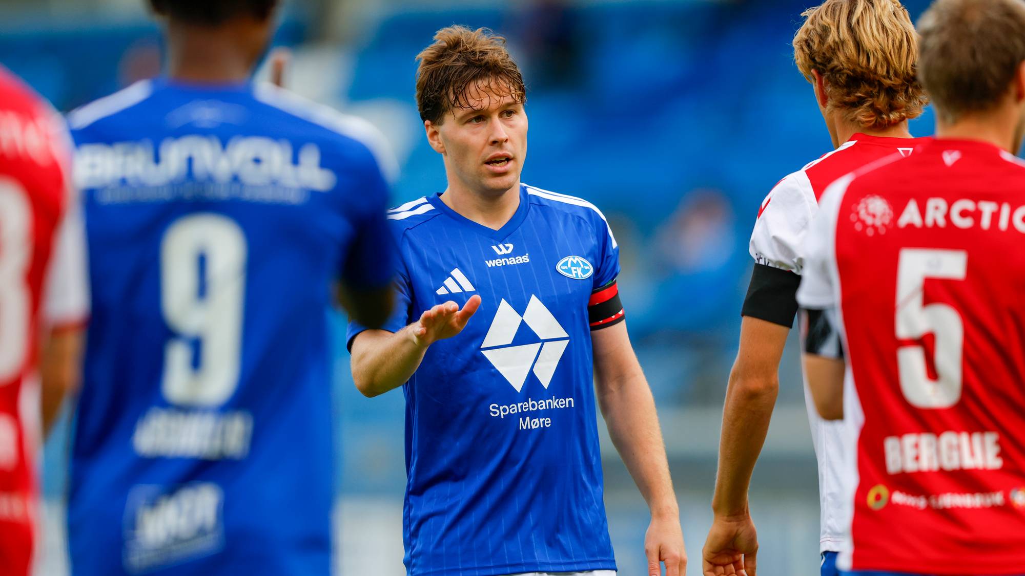 Fotballspiller i blå Molde-drakt gestikulerer til motspillere på banen under en kamp. (Bildebeskrivelsen er laget av en KI-tjeneste)