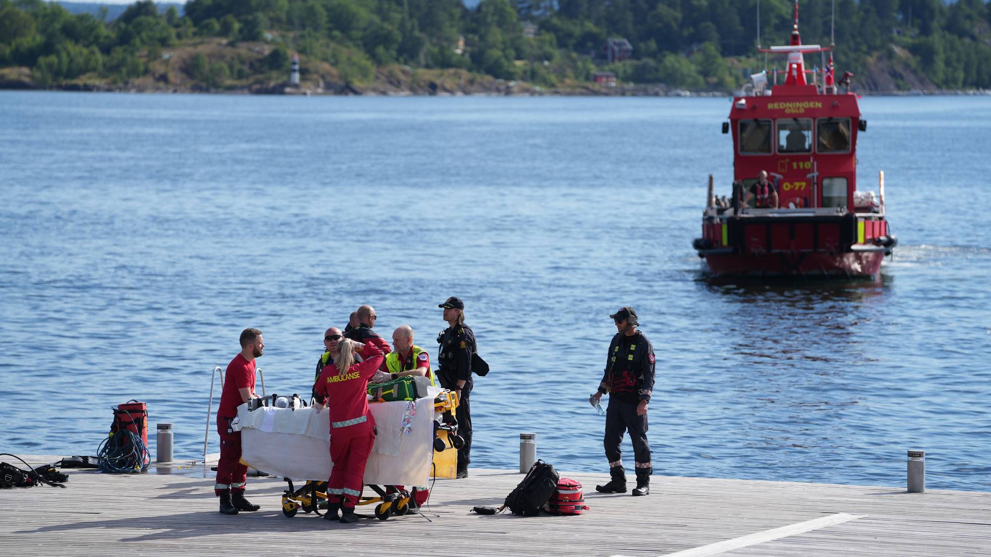 Helsepersonell og politi ved en båre på en brygge med en redningsskøyte i bakgrunnen. (Bildebeskrivelsen er laget av en KI-tjeneste)