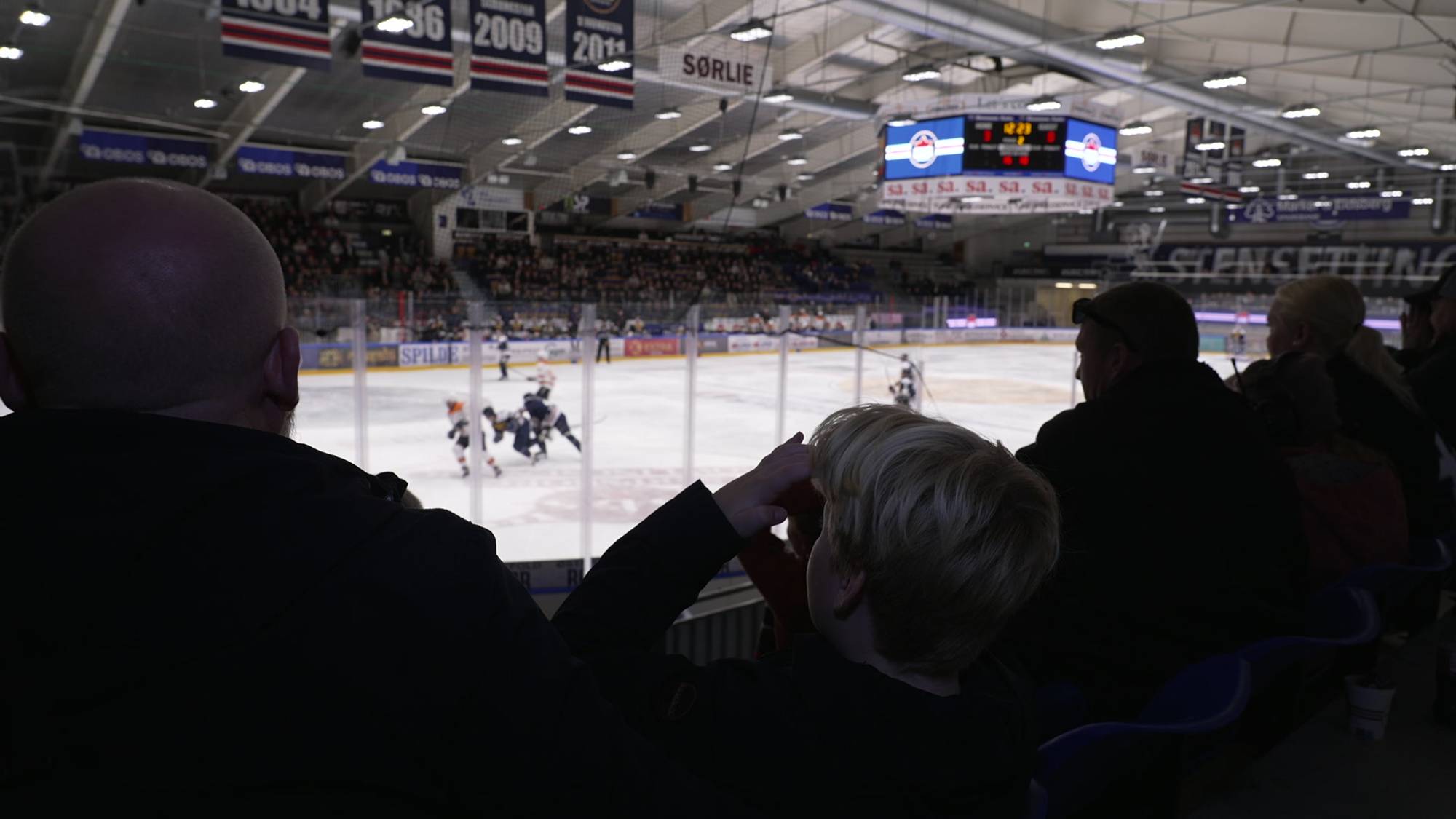 Publikum ser på ishockeykamp fra tribunen i en ishall. (Bildebeskrivelsen er laget av en KI-tjeneste)