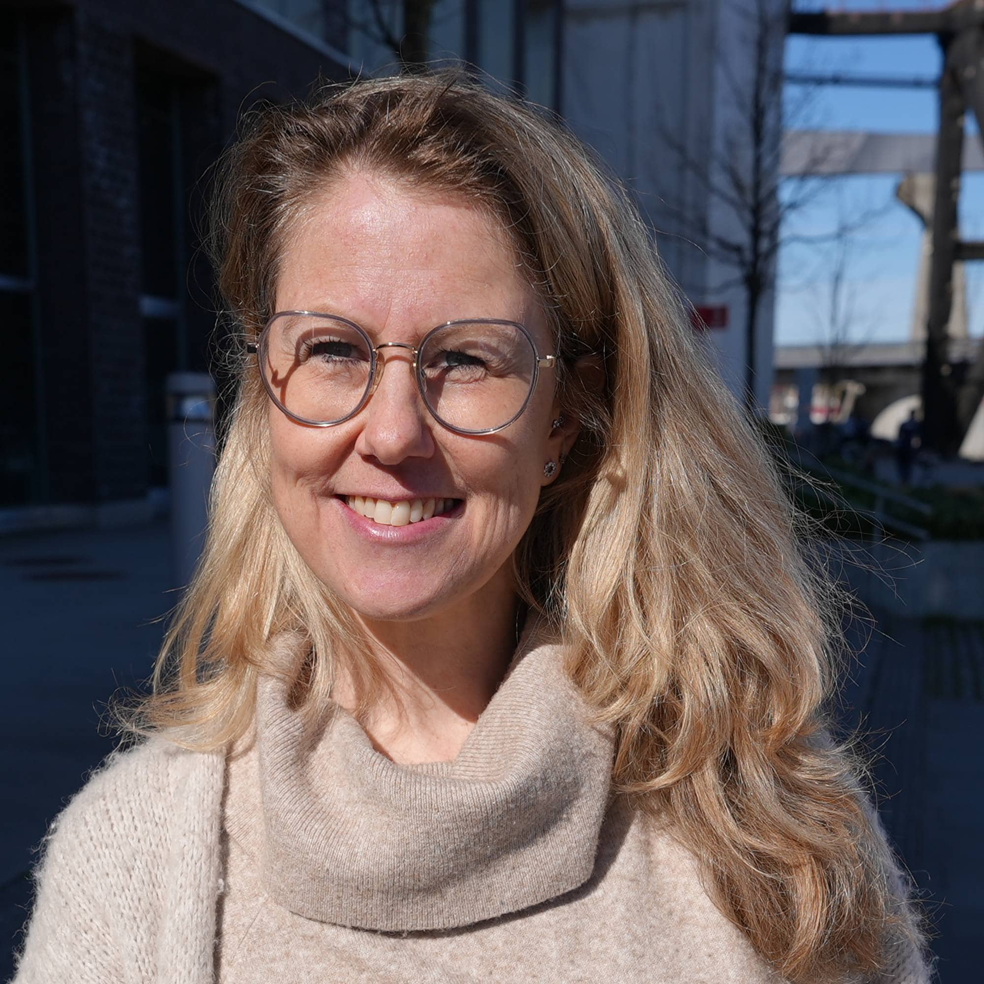 En kvinne med blondt hår og briller står foran en bygning. Hun har på seg en beige genser med høy hals og en cardigan. I bakgrunnen er det en havneinfrastruktur. Sollys kaster lange skygger på bakken. (Bildebeskrivelsen er laget av en KI-tjeneste)