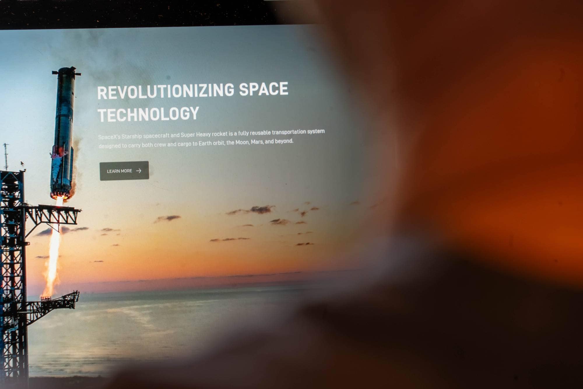 En rakett lyser opp himmelen ved soloppgang. Teksten på bildet sier "REVOLUTIONIZING SPACE TECHNOLOGY". Den beskriver SpaceX's Starship og Super Heavy rocket. Et skjermbilde viser en nettside. (Bildebeskrivelsen er laget av en KI-tjeneste)