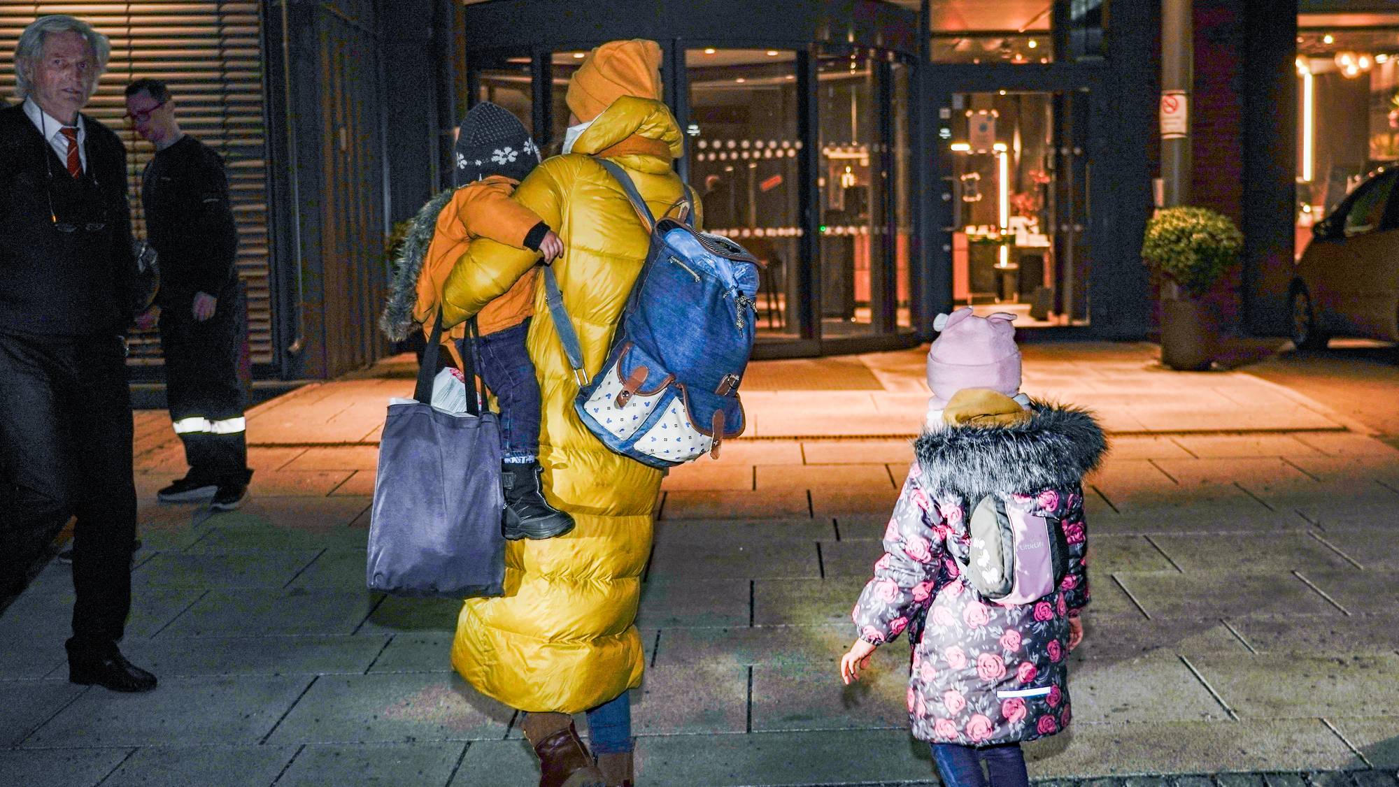 En kvinne med gul jakke bærer et barn, mens en annen jente går ved siden av. Barna er klert i fargerike vinterklær. Bakgrunnen viser inngangen til en bygning med belysning og resepsjon. To personer i uniform overvåker området.