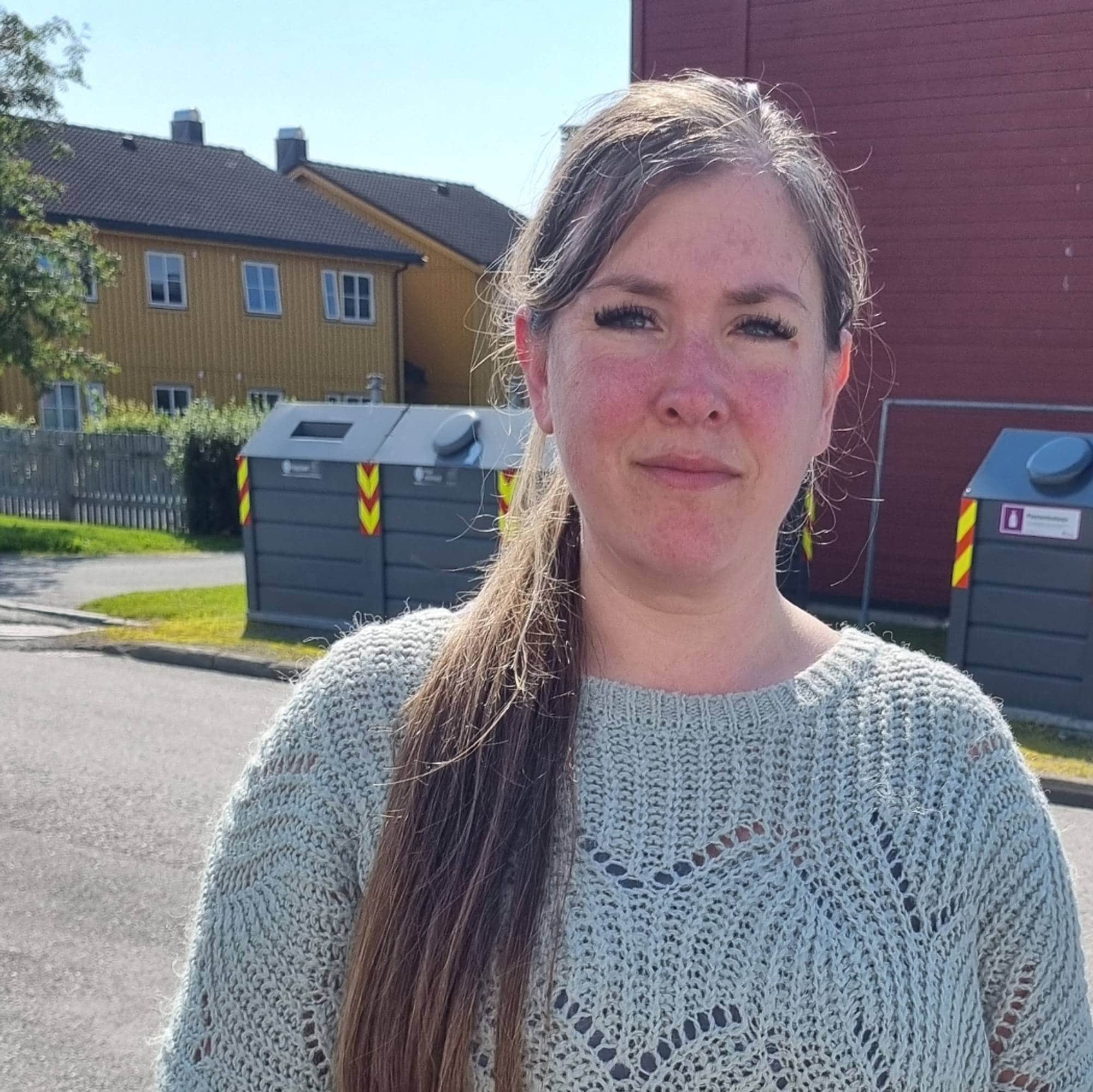 En helfigur av en dame med mørkt langt hår i en hestehale med grå genser som ser inn i kamera. Rødt hus og grusvei i bakgrunnen. 