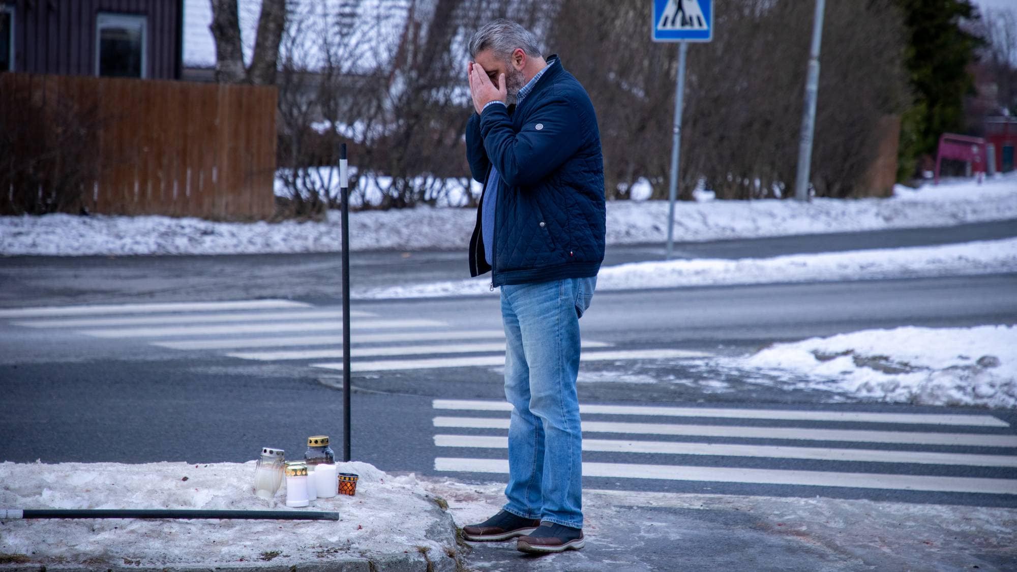 En mann står ved kanten av veien, med hendene i ansiktet. Foran ham er det plassert flere lys på snøen. Området er preget av vinter med snø på bakken og en asfaltvei bak mannen. Det finnes også et trafikkskilt og et gjerde i bakgrunnen.