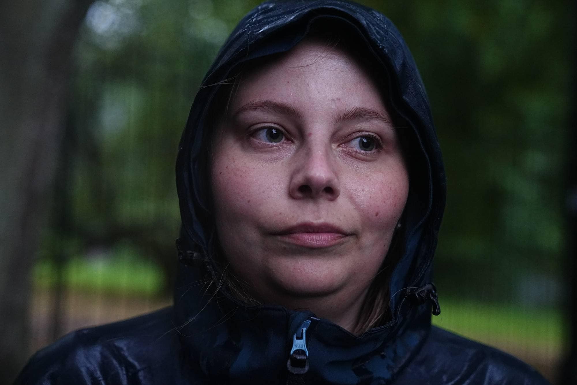 Kvinne i mørk regnjakke med hette oppe ser mot siden i et skogsområde. (Bildebeskrivelsen er laget av en KI-tjeneste)