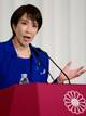 YUICHI YAMAZAKI / Reuters / NTB Sanae Takachi i blå dress gestikulerer mens hun holder en tale ved et podium. Podiet har en rød duk med et blomsterdesign. Det er en vannflaske plassert på podiet. Bakgrunnen er lys og enkel, noe som fokuserer oppmerksomheten på taleren.