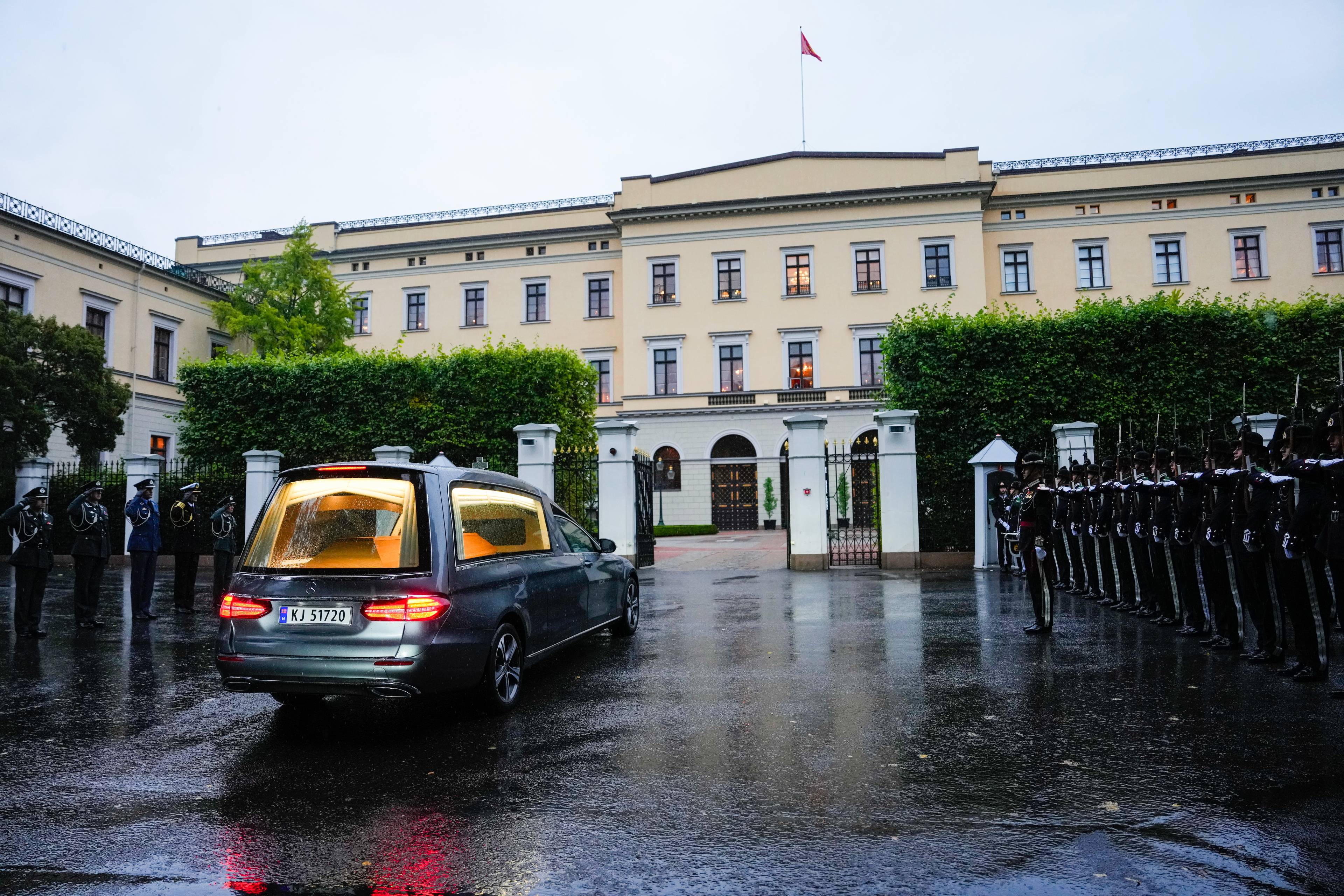 Likbil kjører forbi æresvakt ved Det kongelige slott. Flere uniformerte personer står oppstilt. (Bildebeskrivelsen er laget av en KI-tjeneste)