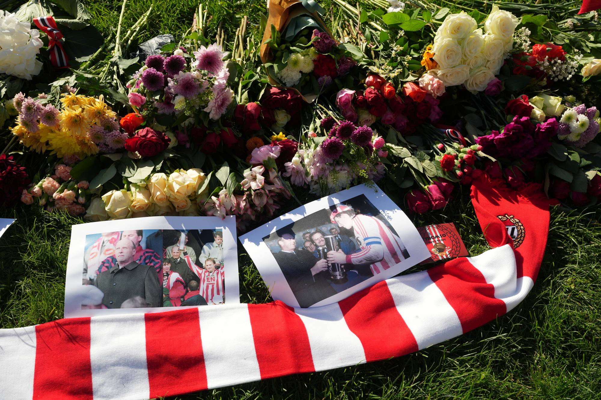 Blomster, fotografier av en fotballspiller og et supporterskjerf ligger på gresset som en minnestund. (Bildebeskrivelsen er laget av en KI-tjeneste)