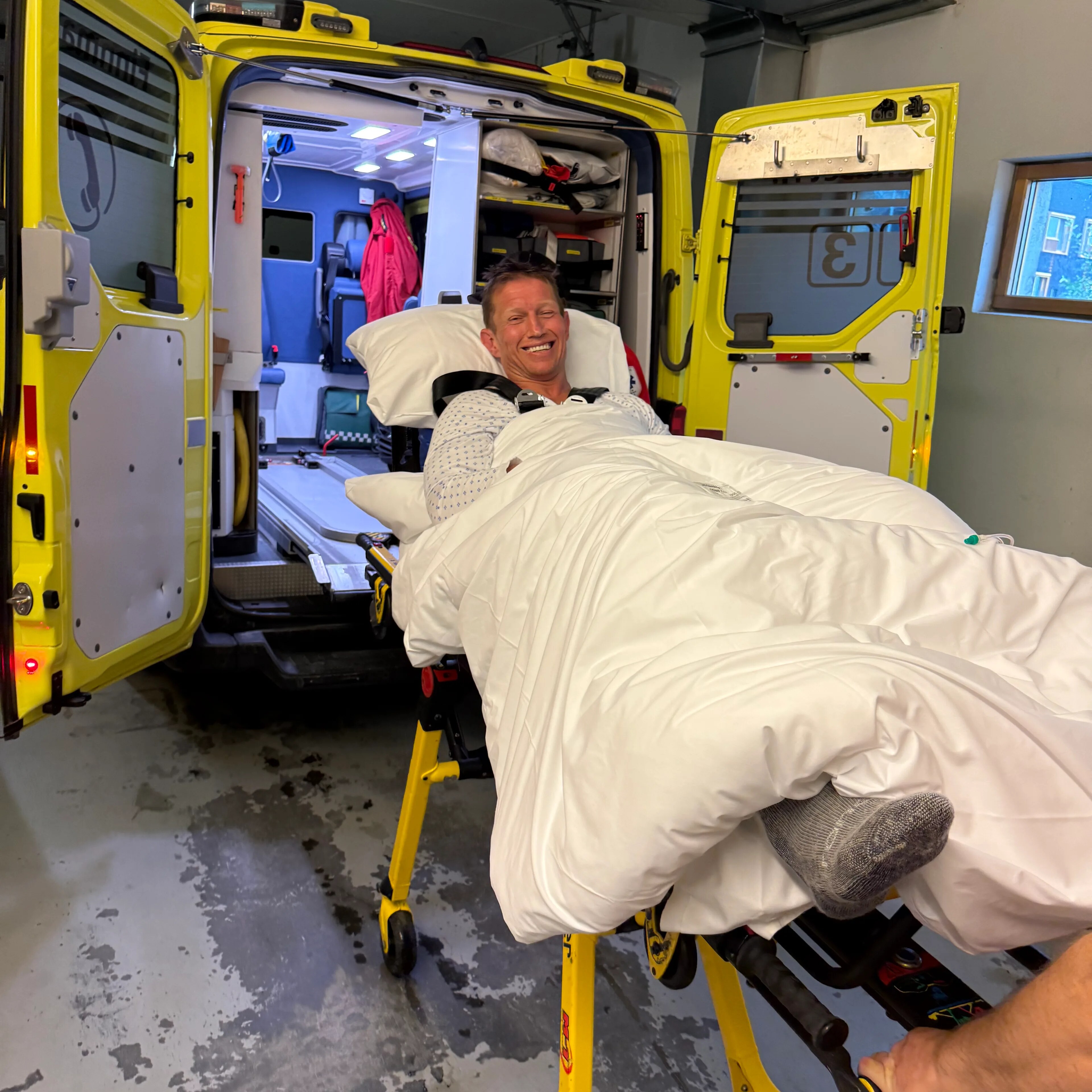 Smilende mann ligger under dyne på en båre utenfor en åpen ambulanse. (Bildebeskrivelsen er laget av en KI-tjeneste)