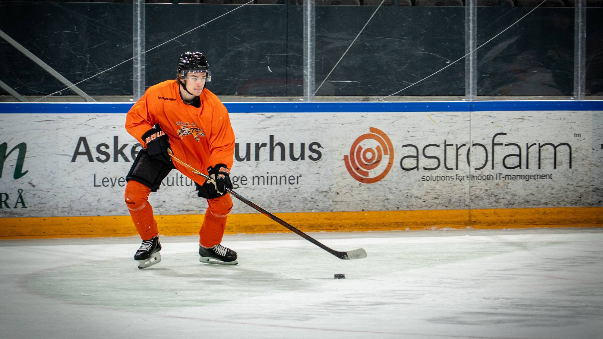 Ishockeyspiller i oransje treningstrøye fører pucken på isen under en økt i en ishall. (Bildebeskrivelsen er laget av en KI-tjeneste)