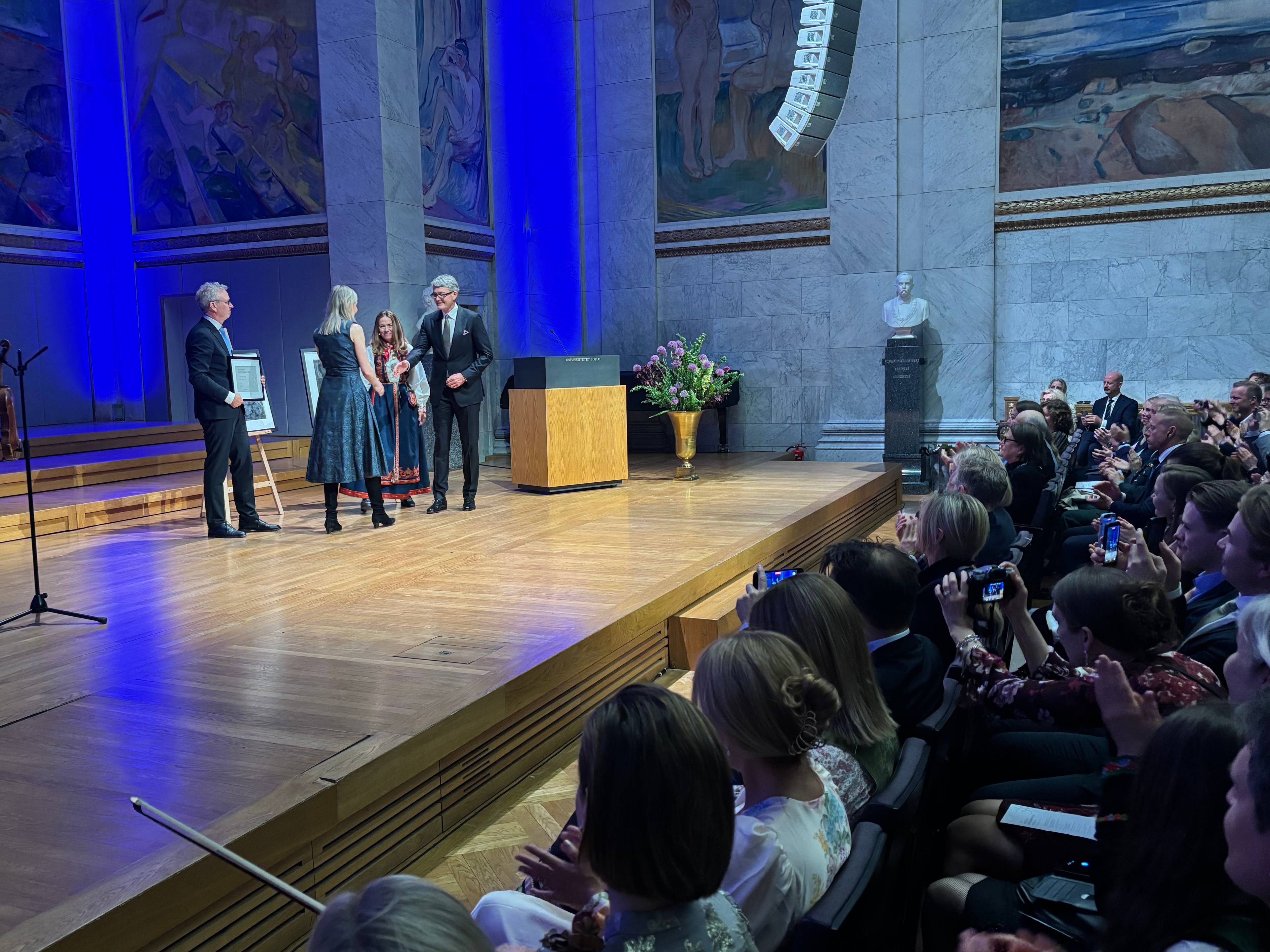 fire menneske står på ei scene, og ei mengde publikum sit i salen føre scena