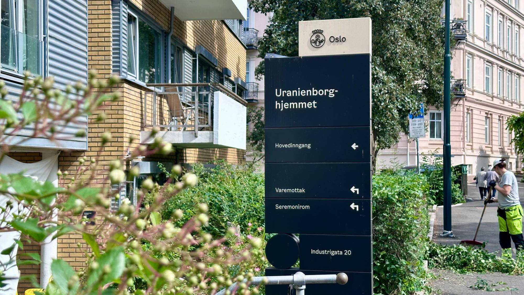 Et skilt viser vei til Uranienborg-hjemmet, hovedinngang, varemottak og seremonirum. En ansatt raker opp løv på en fortau. Bygninger og grøntområder utgjør bakgrunnen. (Bildebeskrivelsen er laget av en KI-tjeneste)