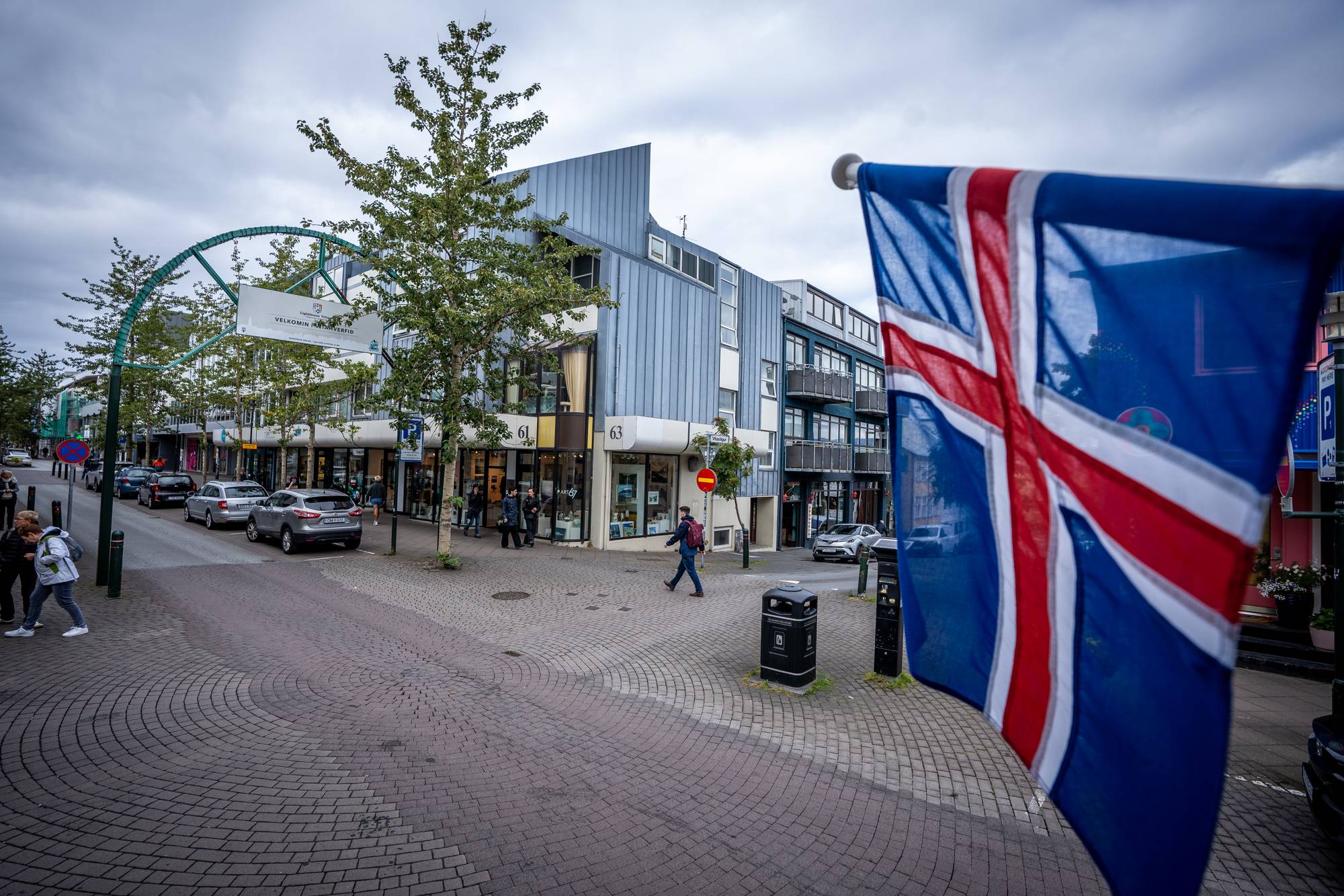 Islandsk flagg i forgrunnen foran bygningene i en gågate i Reykjavik. Folk går i gaten. (Bildebeskrivelsen er laget av en KI-tjeneste)