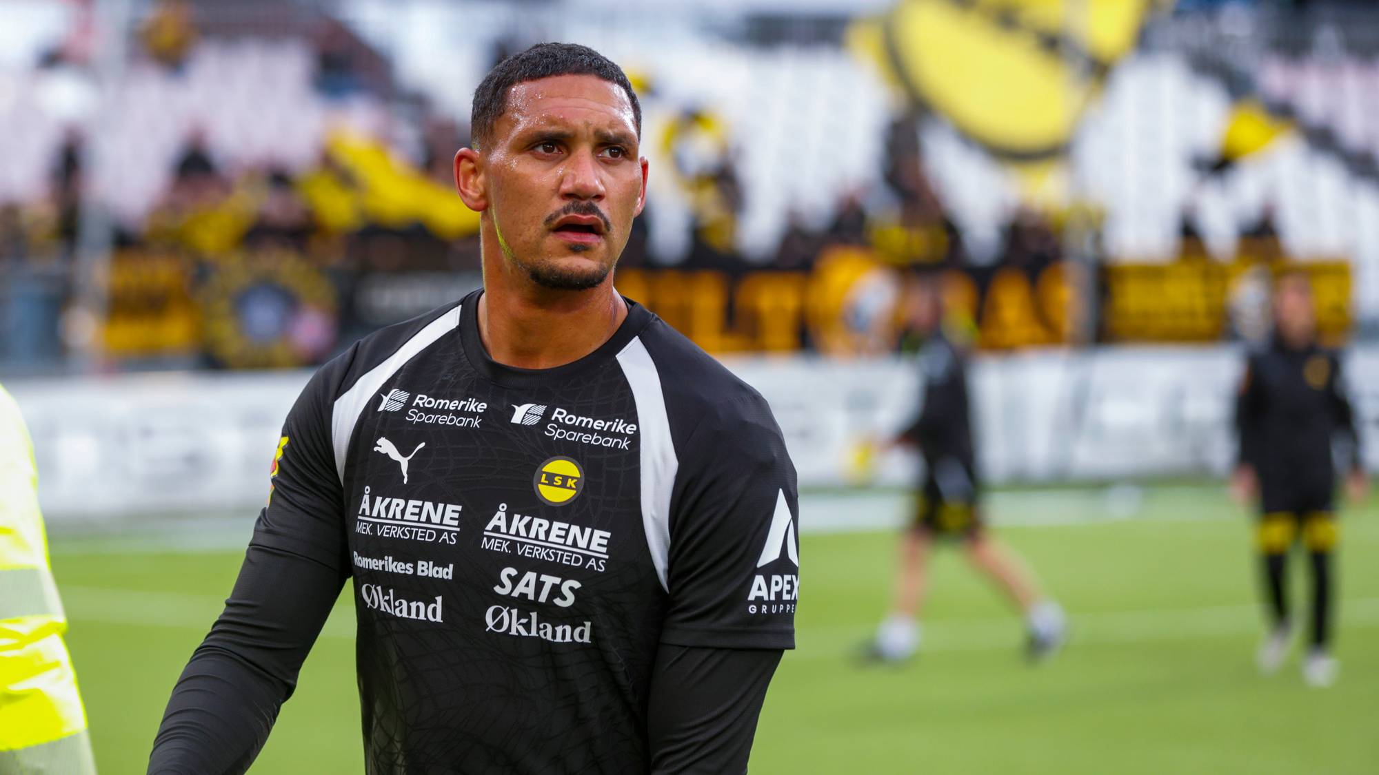 En fotballspiller i svart drakt ser fremover på en gressbane. Bildet er tatt utendørs, trolig under en kamp. Spilleren har en logo fra LSK på brystet. I bakgrunnen er det andre personer og tribuner. (Bildebeskrivelsen er laget av en KI-tjeneste)