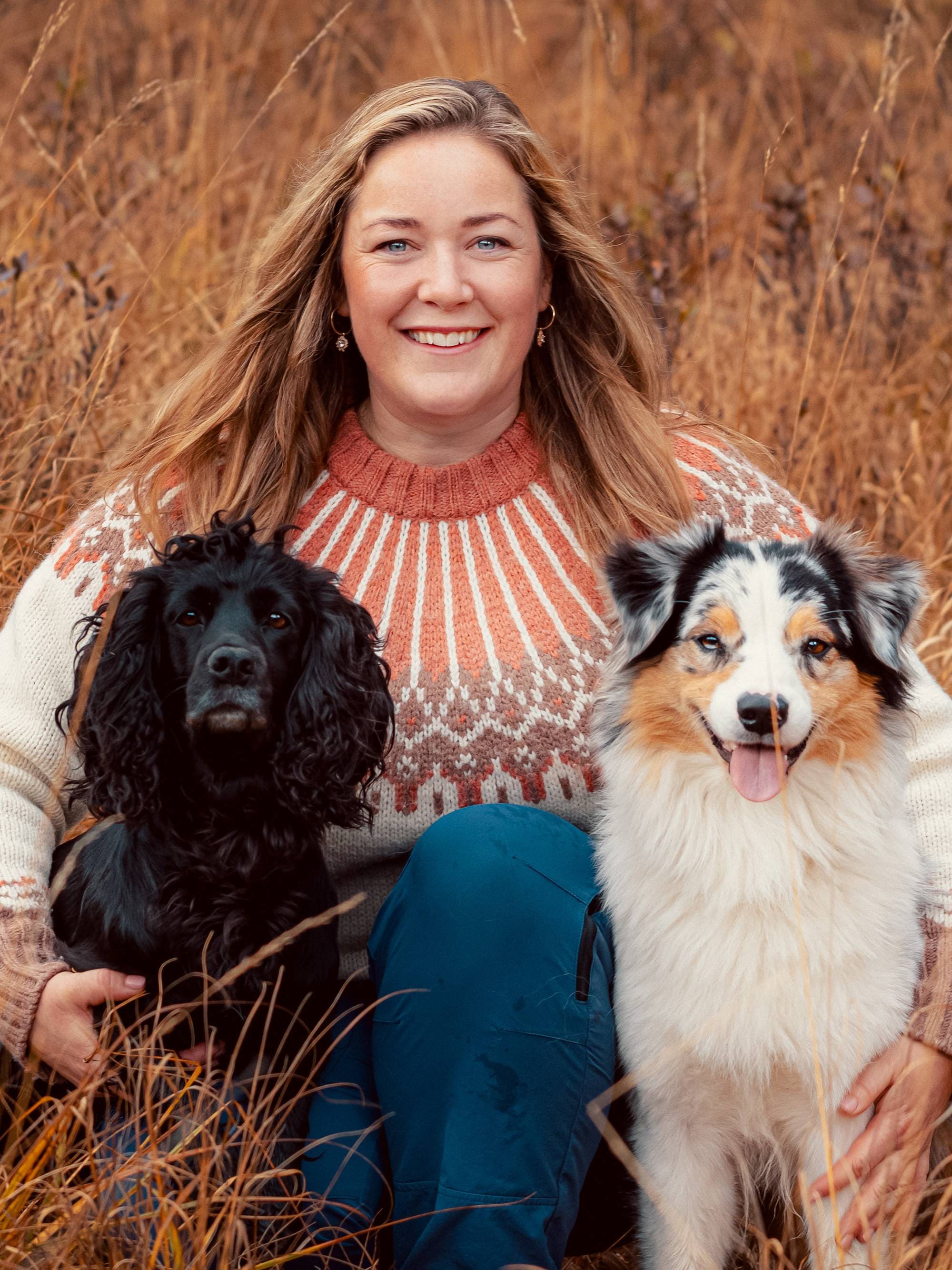 Hundetrener Maren Teien Rørvik sitter på bakken. Hun har en hund i hver armkrok (sine hunder). Den ene er en svart cocker spaniel, den andre en spraglete gjeterhund. Maren har langt, brunt hår. Hun har på seg olabukse og ullgenser med mønster i oransje og beige. Hun smiler. 