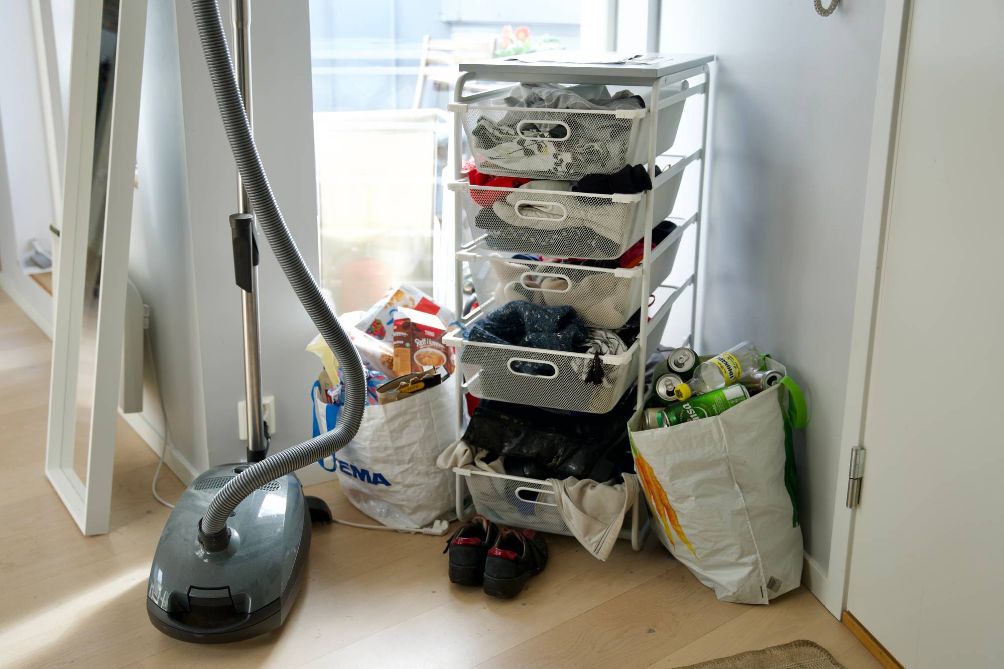 I hjørnet av stua i kollektivets leilighet, står det en hylle til oppbevaring av klær og tilbehør, en pose til pant, en pose til papp-søppel og en støvsuger. 