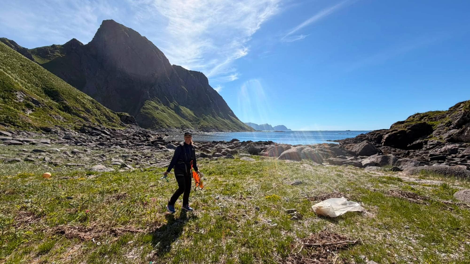 En dame i sort tøy med caps på hode rydder søppel på en gresslette like ved kysten av Mosken. I bakrunn ser man høye, grønne, spisse fjell og hav.