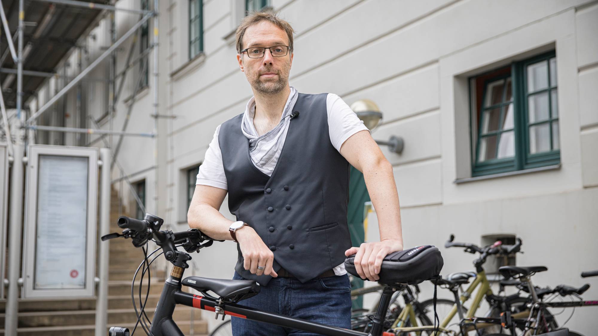 En mann med briller står utendørs og støtter seg til en sykkel foran en lys bygningsfasade. (Bildebeskrivelsen er laget av en KI-tjeneste)