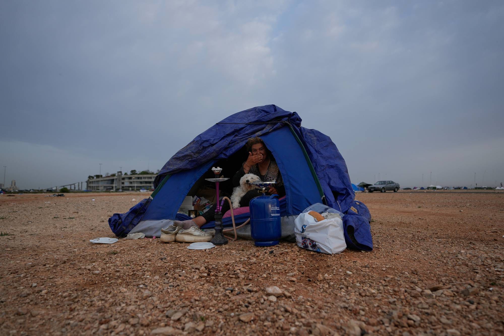 En kvinne og en hund sitter inne i et blått telt i et øde landskap. Utenfor teltet ligger diverse gjenstander. I bakgrunnen kan man se en bygning, biler og andre telt. (Bildebeskrivelsen er laget av en KI-tjeneste)