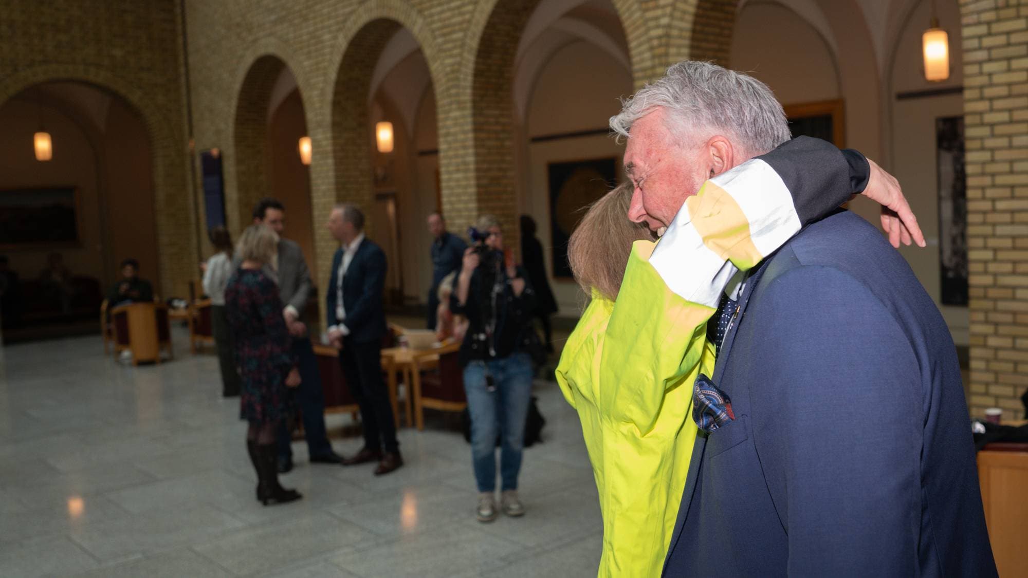 En eldre mann i mørk dress blir omfavnet av en person i en knallgul jakke. Bak dem står andre personer og en fotograf med kamera. Bygningen har buede ganger og mursteinsvegger. (Bildebeskrivelsen er laget av en KI-tjeneste)