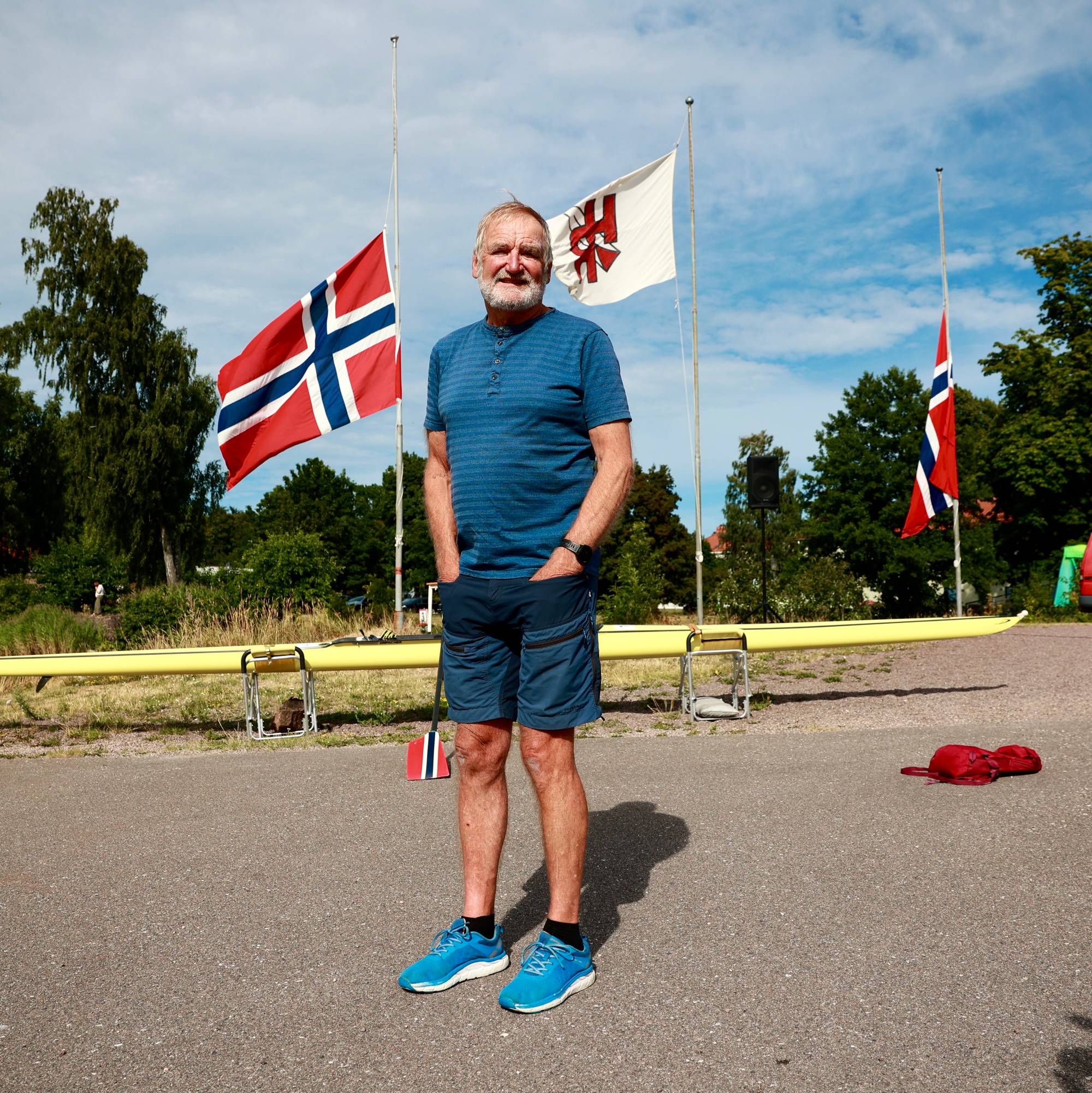 En eldre mann står utendørs foran en robåt, norske flagg og trær.