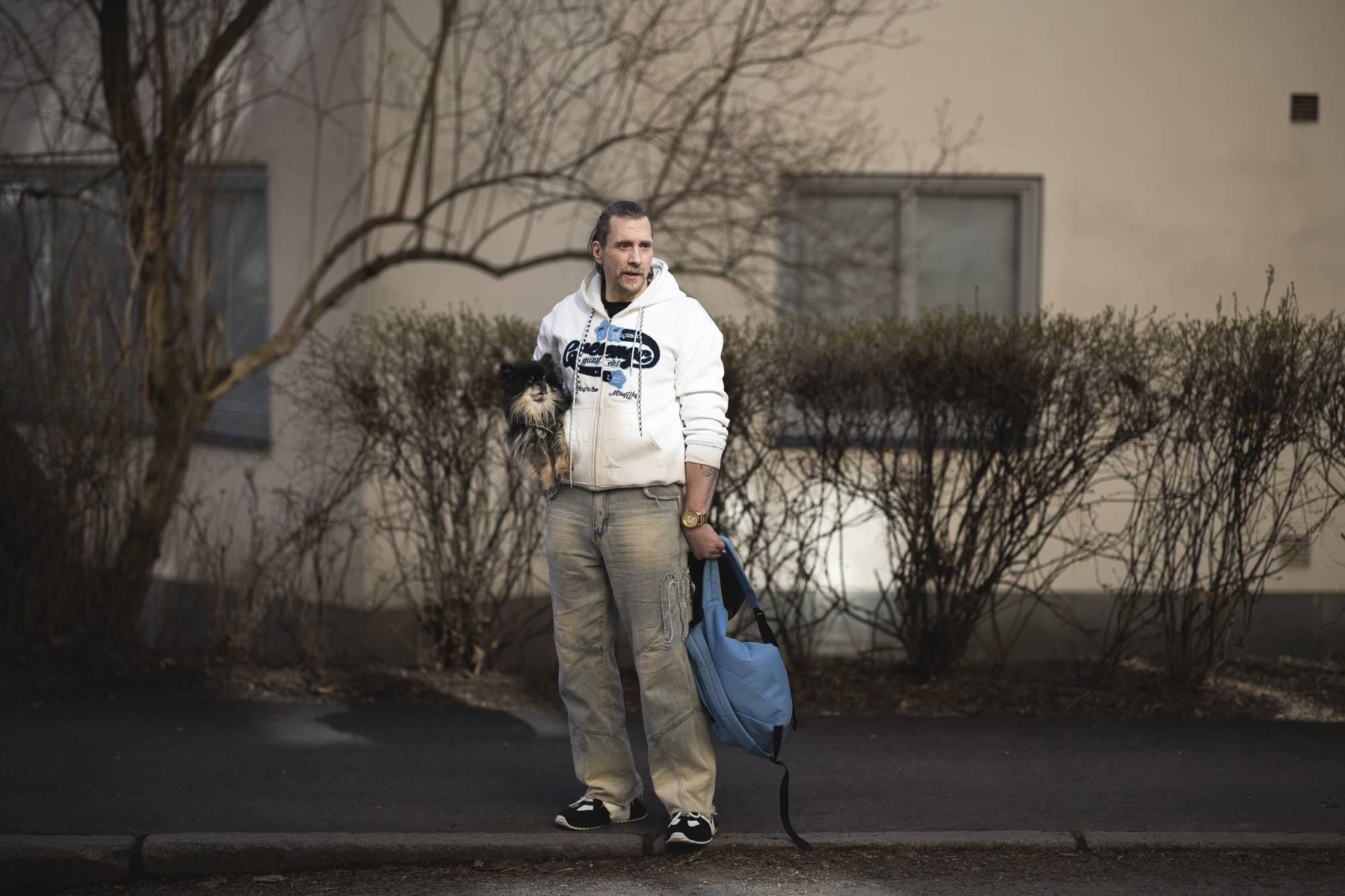 En mann holder en liten hund i armene. Han står utenfor en bygning med en lyseblå bag i den andre hånden. Mannen har på seg en hvit hettegenser og jeans. Busker og trær rammer inn scenen. (Bildebeskrivelsen er laget av en KI-tjeneste)