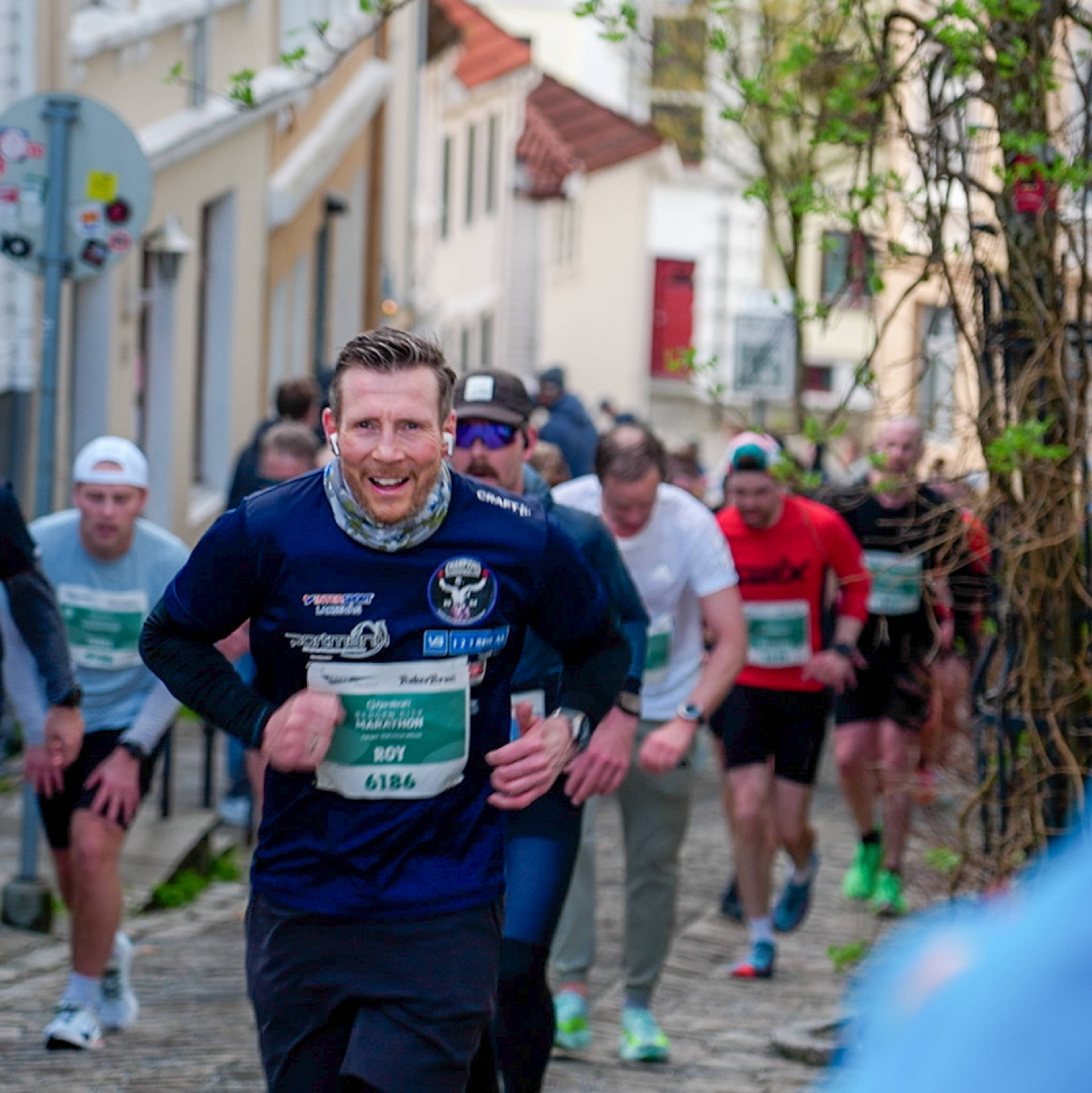 Løparar spring mot kamera opp ei smal gate på Nordnes under Bergen City Marathon, med kvite trehus i sollys i bakgrunnen.