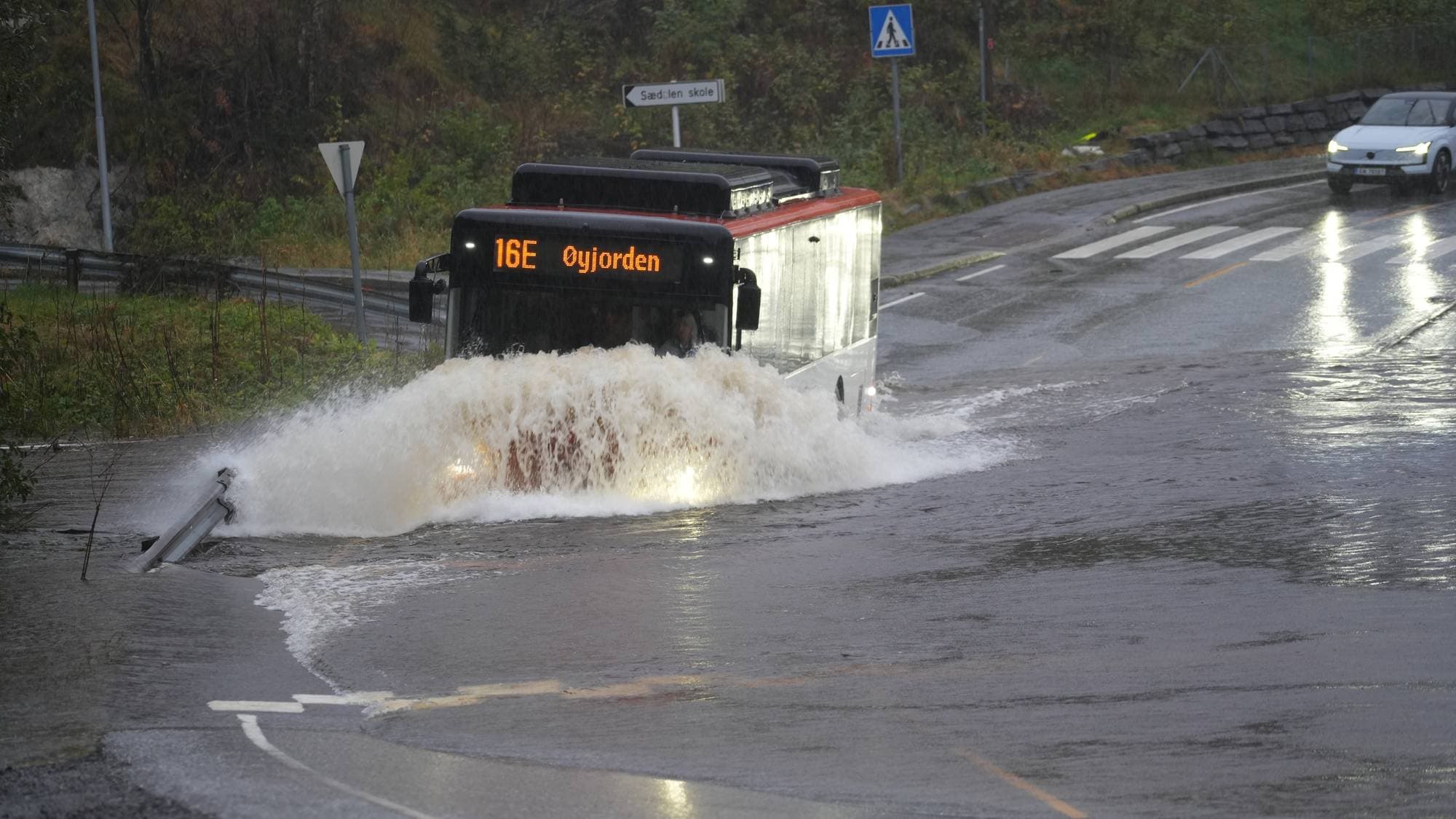 Buss kjører gjennom en oversvømt vei. Vannet spruter opp foran kjøretøyet. En bil venter i bakgrunnen. (Bildebeskrivelsen er laget av en KI-tjeneste)
