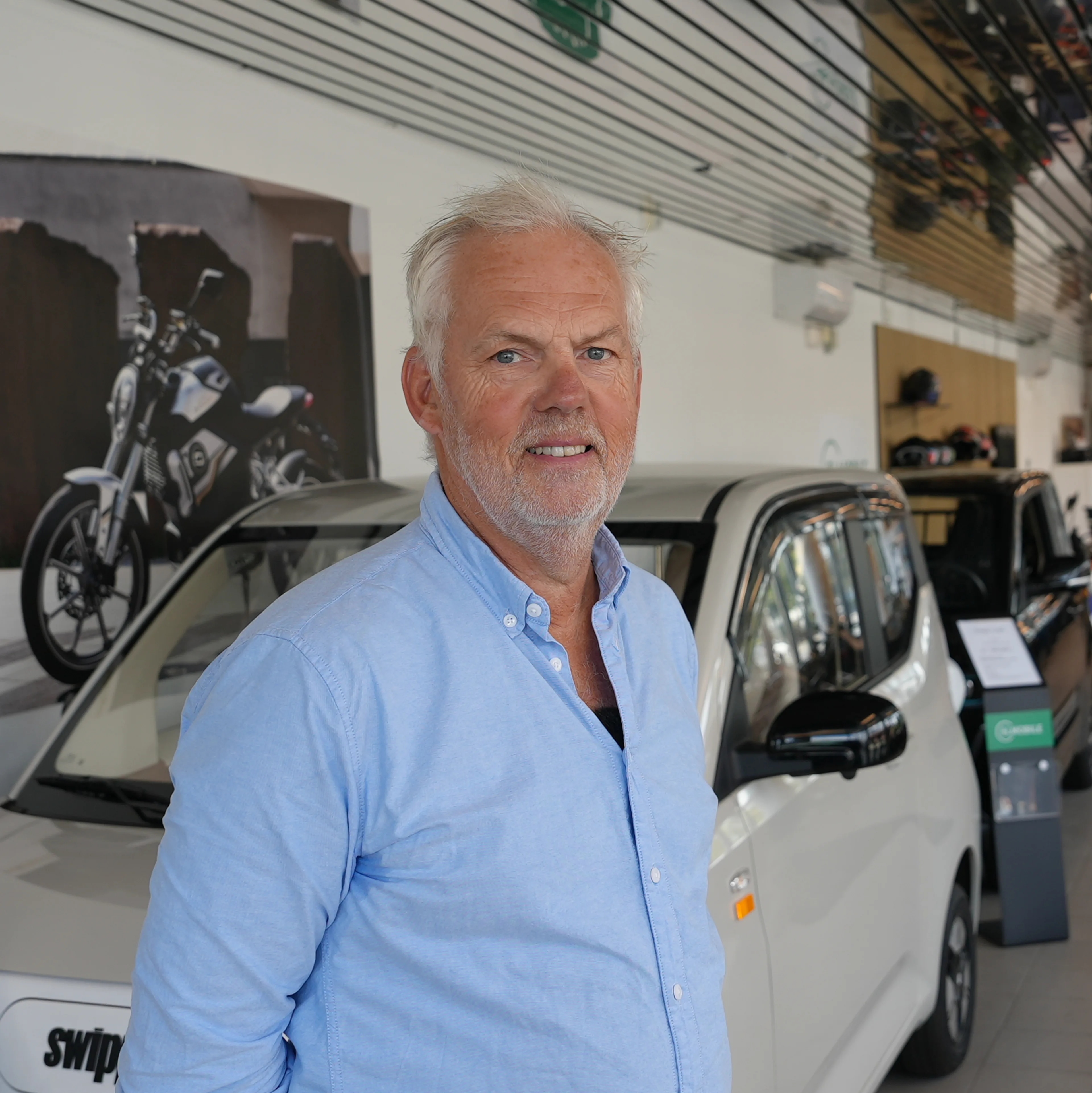 Olav står foran og i mopedbilen og viser oss hvordan den fungerer. Han smiler og har på seg en blå skjorte.