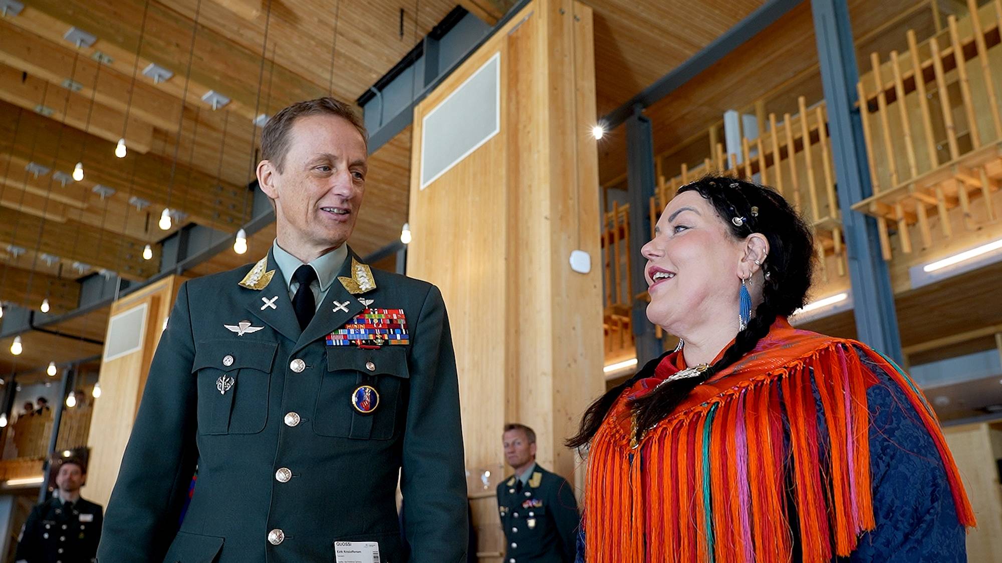 Mann i militæruniform snakker med en kvinne i tradisjonell samisk kofte. Bak dem står flere andre personer i militæruniform.