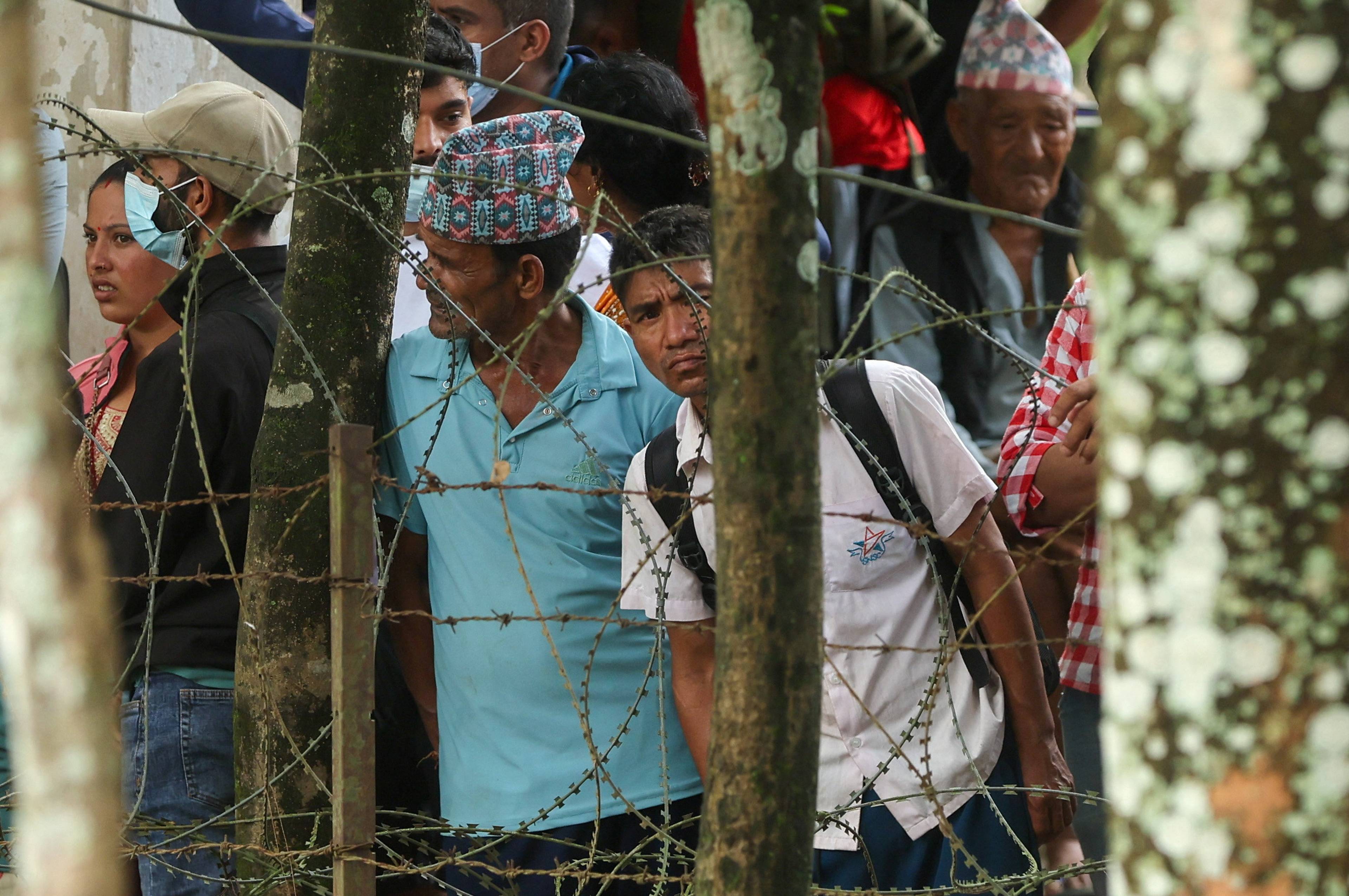 Mennesker står bak et piggtrådgjerde. En mann har på seg en tradisjonell nepalsk lue. (Bildebeskrivelsen er laget av en KI-tjeneste)