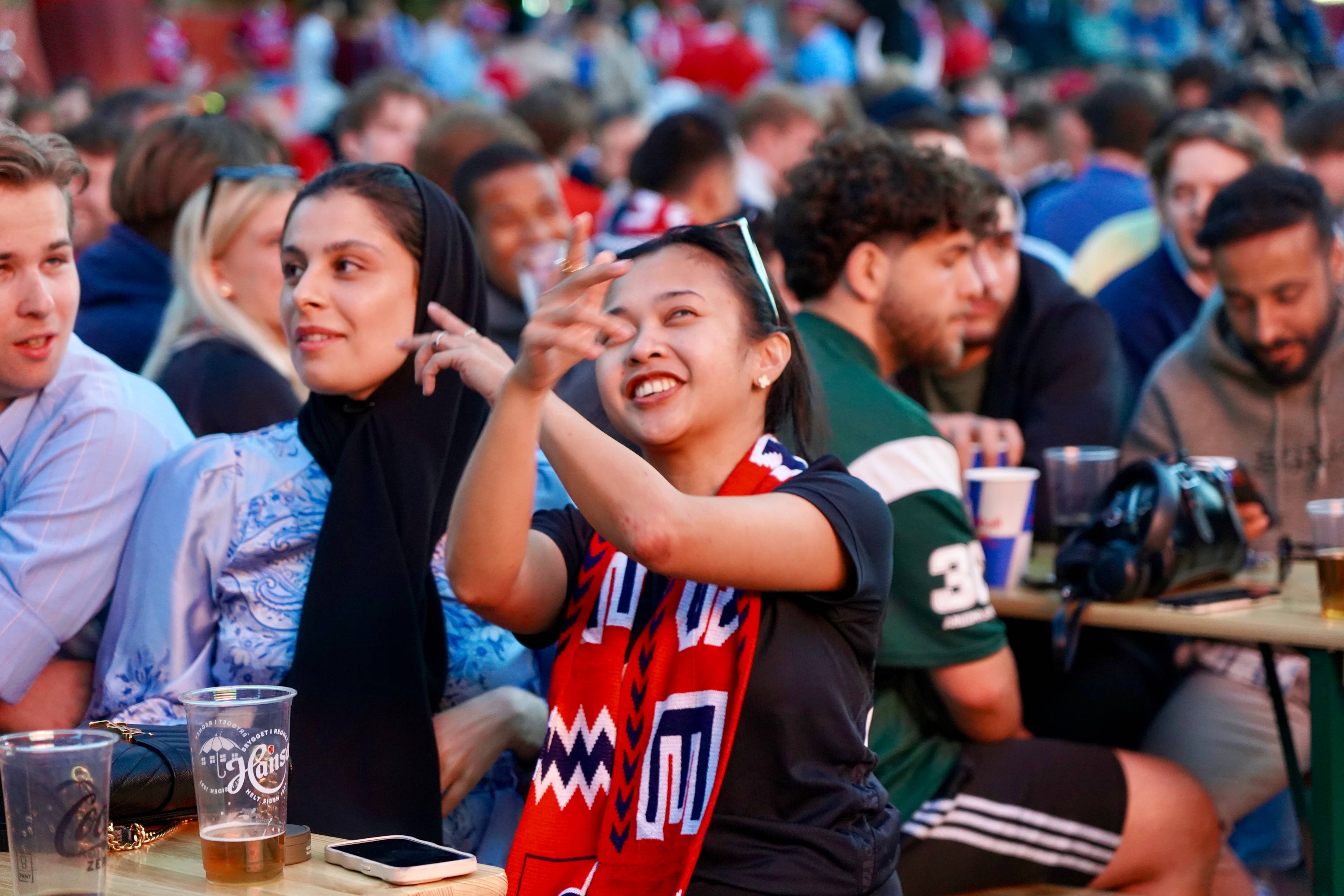 En kvinne i forgrunnen vinker til noen mens hun har på seg et supporter-skjerf. Bak henne sitter en gruppe mennesker tett samlet. De sitter ved et bord med drikkevarer foran seg. Det er tydelig at de er tilskuere.