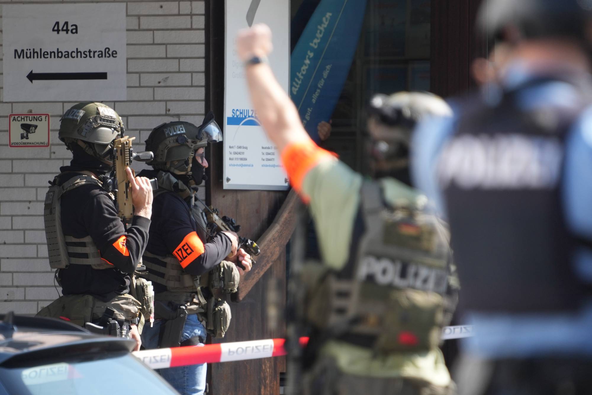 Tunge bevæpnede politifolk i fullt utstyr er synlige utenfor en bygning. En politimann holder et automatgevær, mens en annen ser til siden med en pistol i hånden. De er på et sted som er sperret av med polititape.