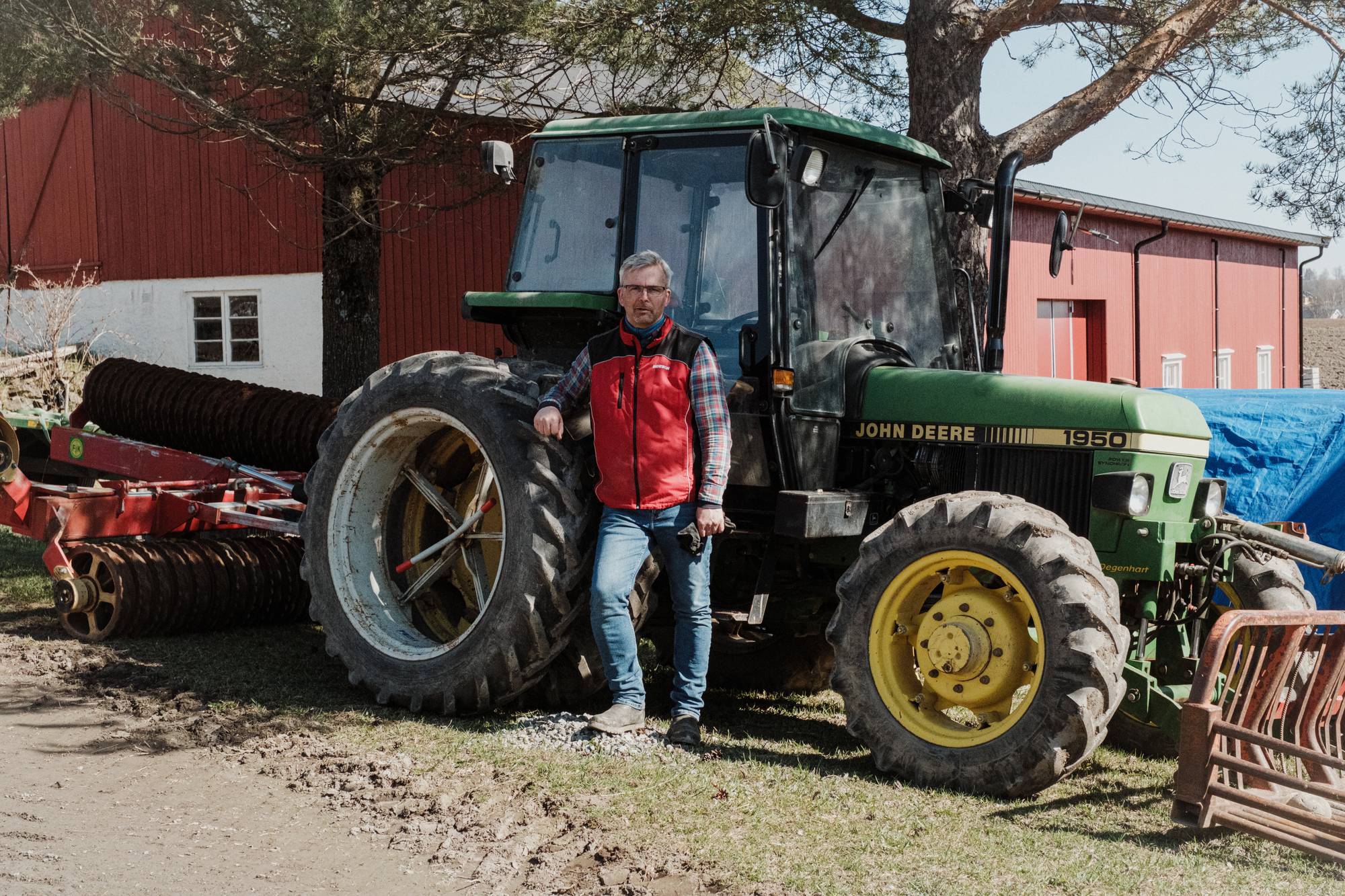 Bror Helgestad, en mann i rutete skjorte, rød boblevest og jeans lener seg mot en traktor og ser i kamera