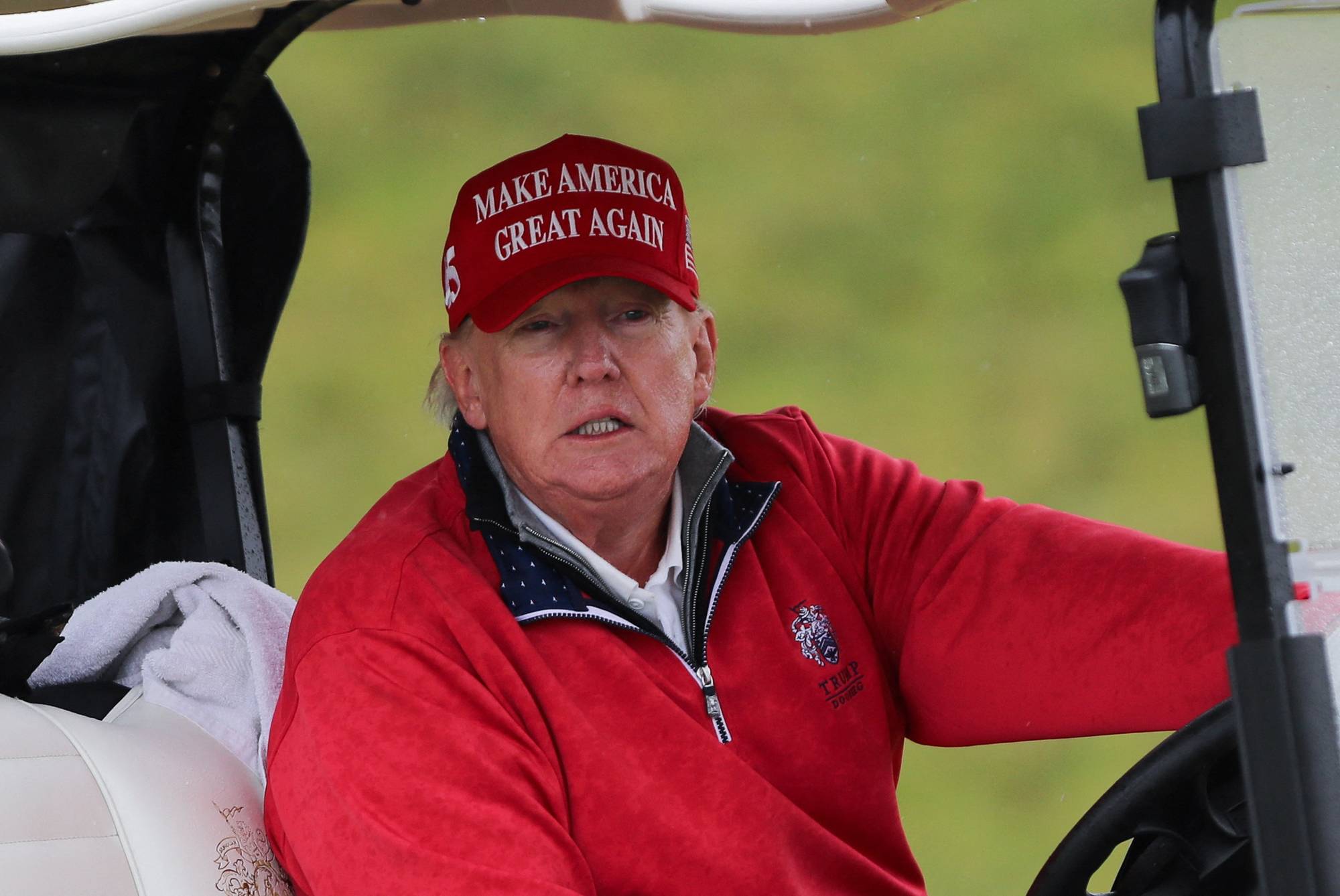 Donald Trump sitter i en golfbil iført rød caps med tekst og rød genser. (Bildebeskrivelsen er laget av en KI-tjeneste)