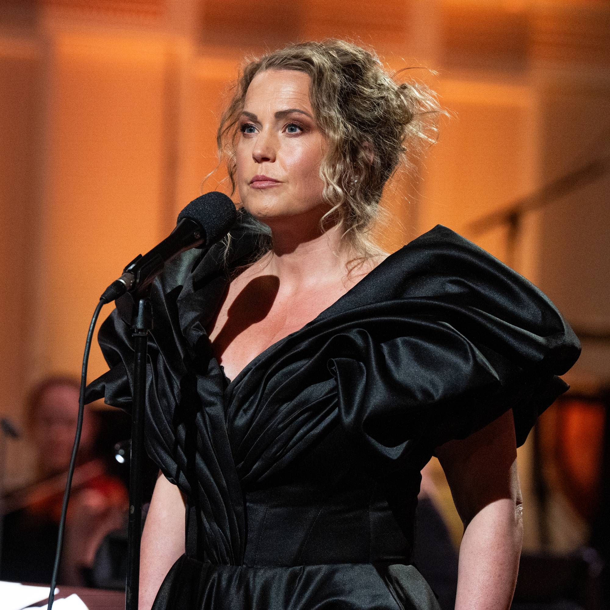 En kvinne i svart kjole synger i en mikrofon under en konsert. En dirigent leder et orkester i bakgrunnen. Bakgrunnen er opplyst med oransje lys. (Bildebeskrivelsen er laget av en KI-tjeneste)
