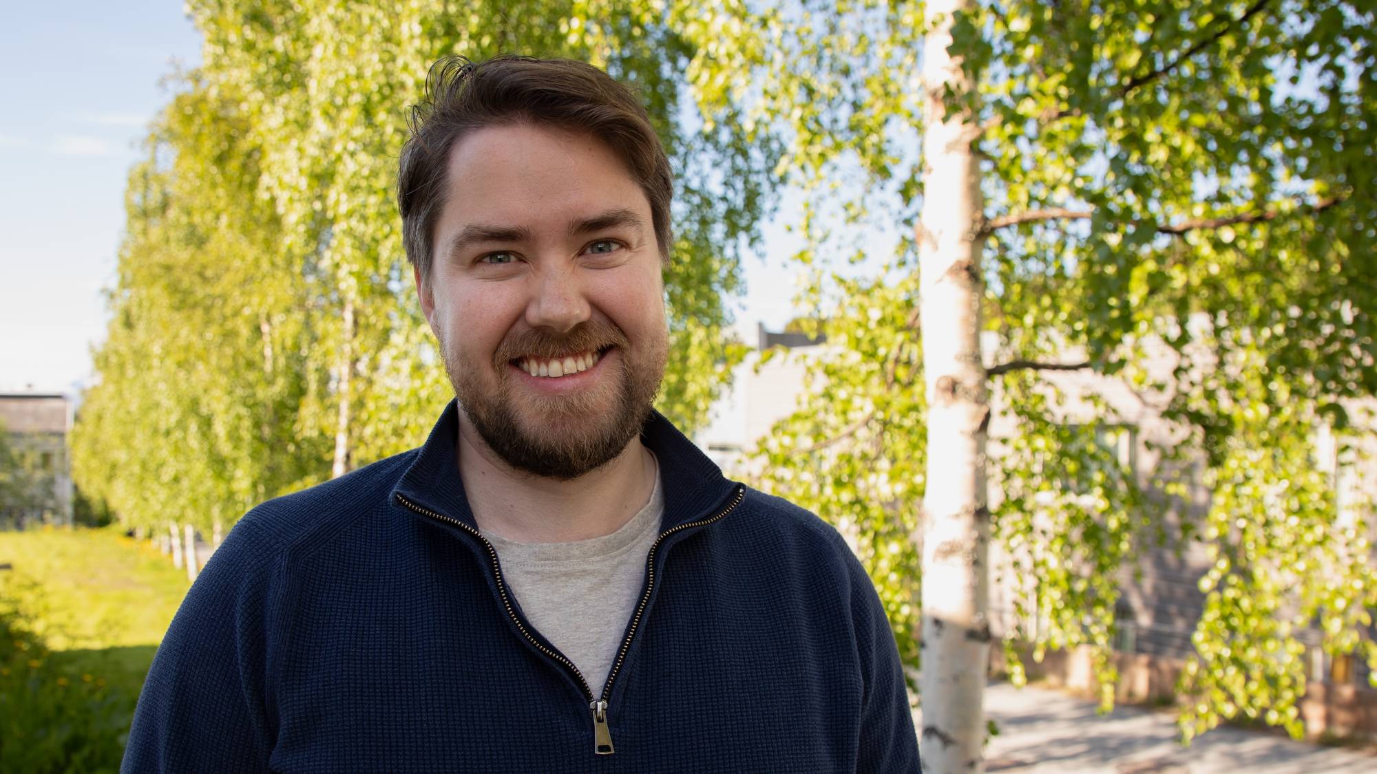 En mann med brunt hår i half zip smiler foran et grønt landskap. Halvnært portrett.