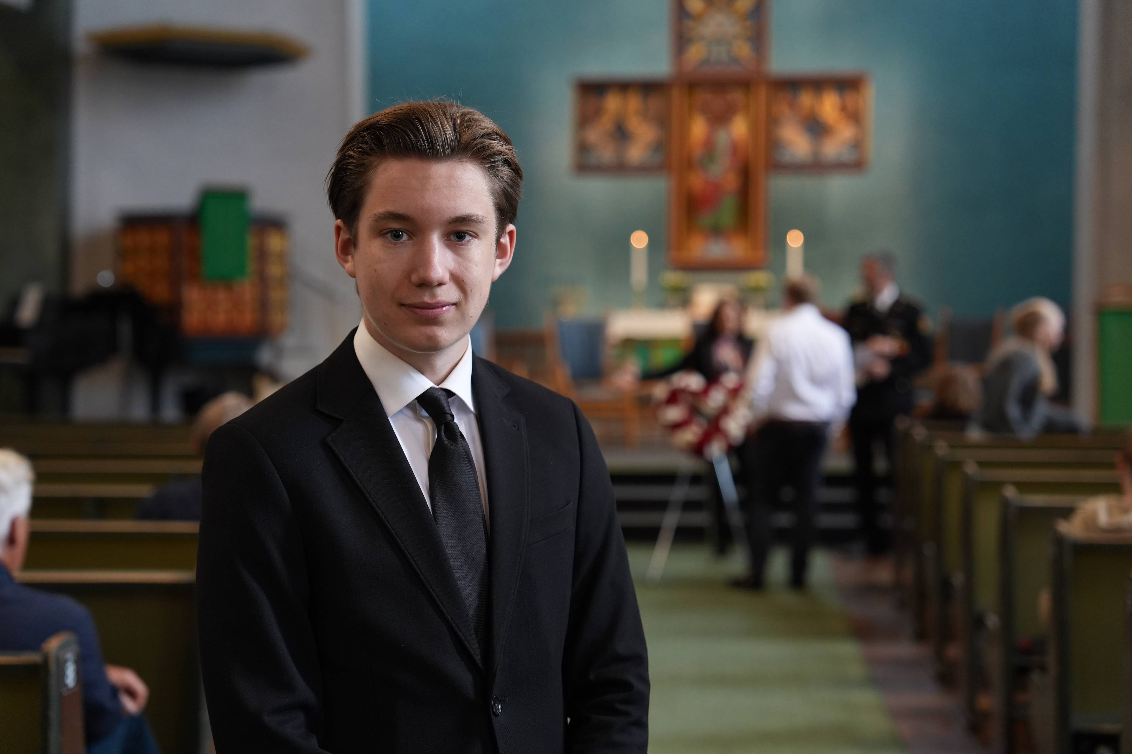 Ung mann i dress i en kirke, med andre mennesker og et kors i bakgrunnen. (Bildebeskrivelsen er laget av en KI-tjeneste)