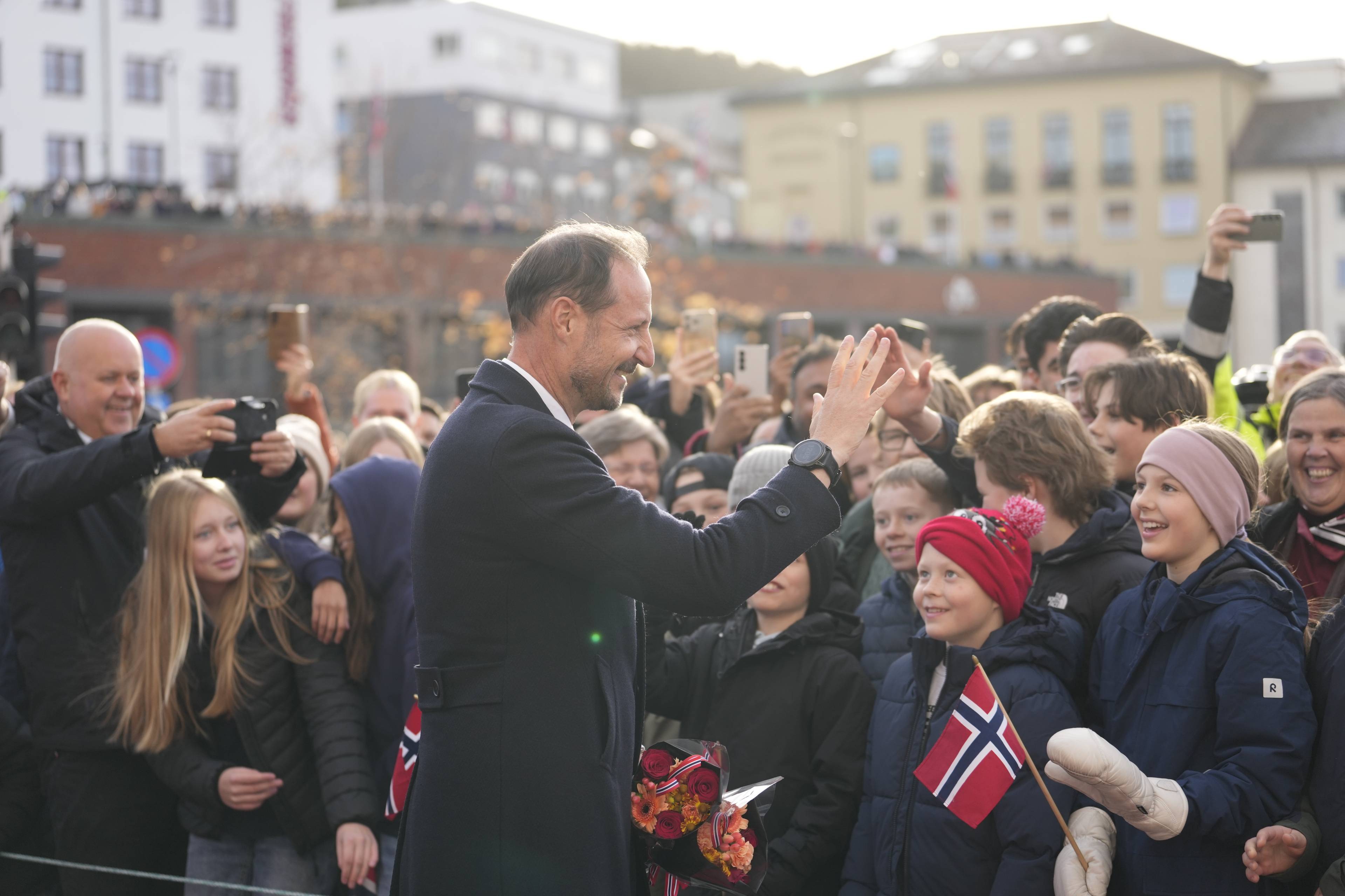 Kronprins Haakon hilser på folkemengden utendørs i bymiljø. (Bildebeskrivelsen er laget av en KI-tjeneste)