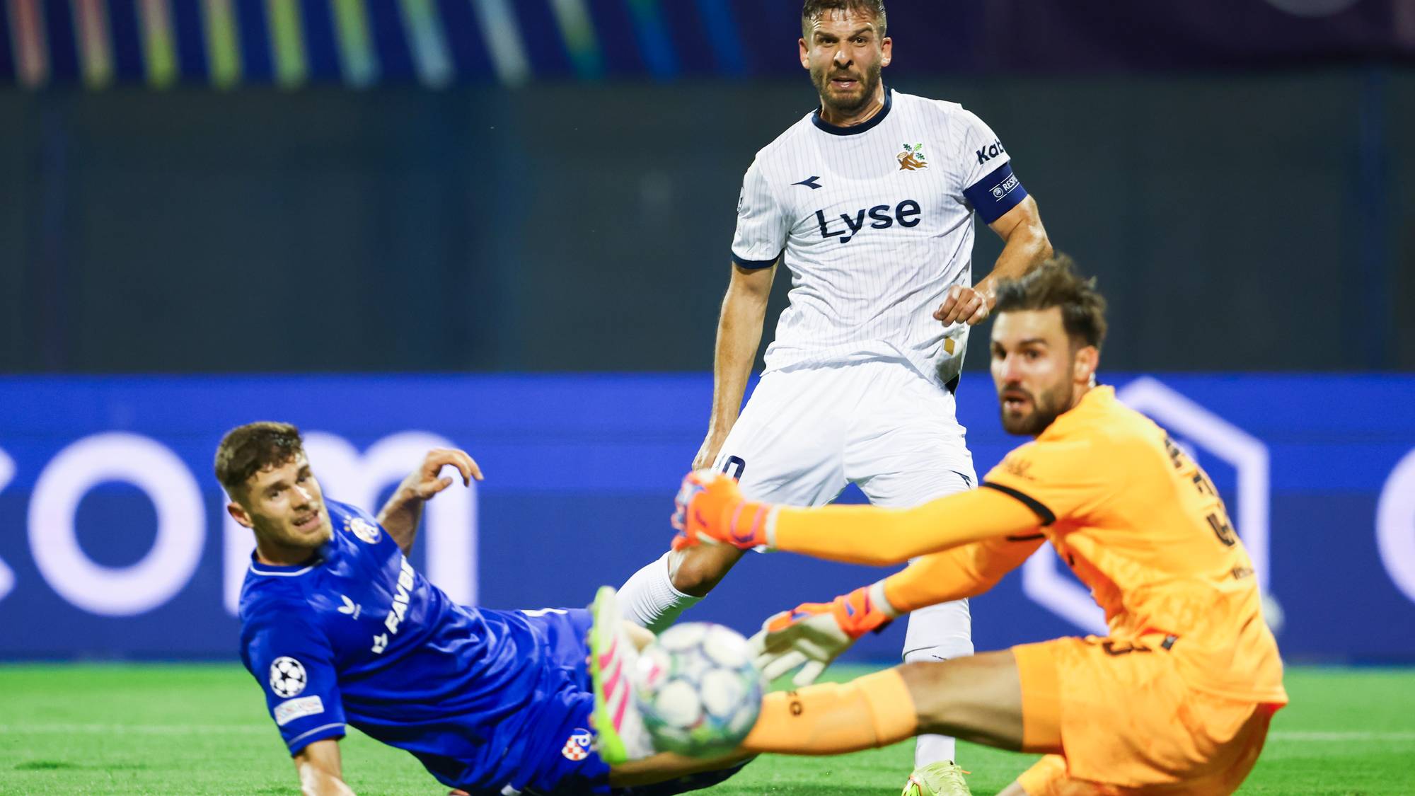 Fotballspiller i blå drakt prøver å nå ballen mens keeper i oransje redder, med spiller i hvitt bak. (Bildebeskrivelsen er laget av en KI-tjeneste)