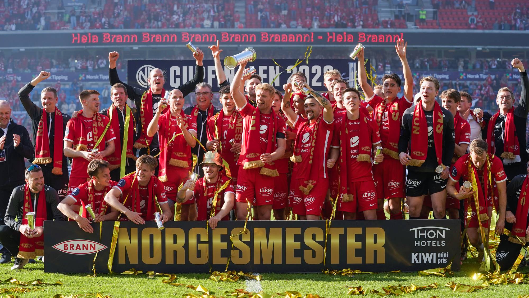 Fotballaget feirer med troféer og konfetti. De står bak et banner med teksten "NORGMESTER". En stor folkemengde i rødt sitter på tribunen. (Bildebeskrivelsen er laget av en KI-tjeneste)