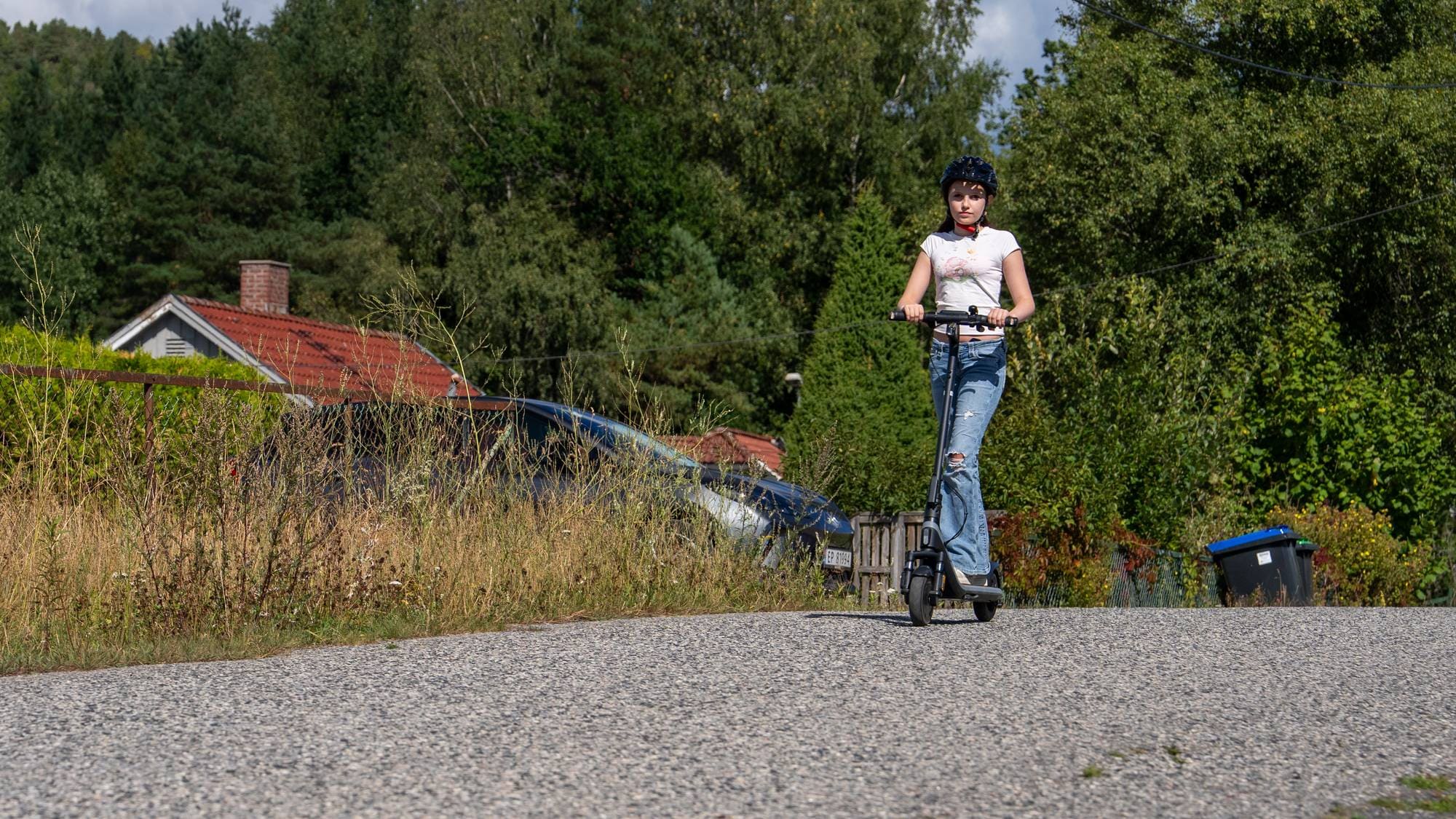 Ung jente med hjelm på elsparkesykkel på asfaltvei i boligstrøk. 