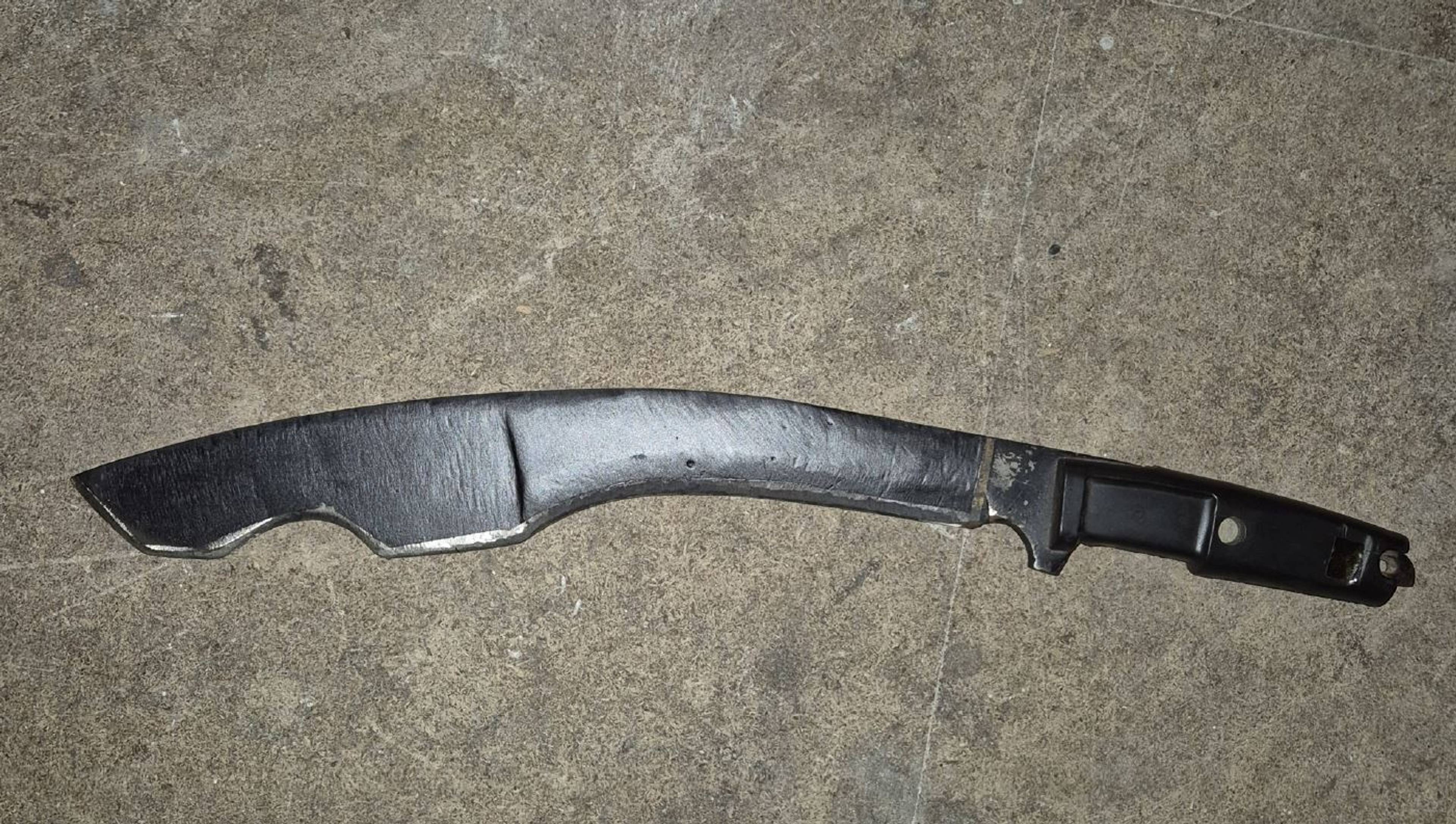 En stort, buet machete med et svart håndtak ligger på et gulv eller bord. Macheten har en tykk, matt metallklinge. Klingen er bredere mot spissen. Håndtaket ser ut til å være av plast og har en rektangulær utforming med avrundede hjørner.