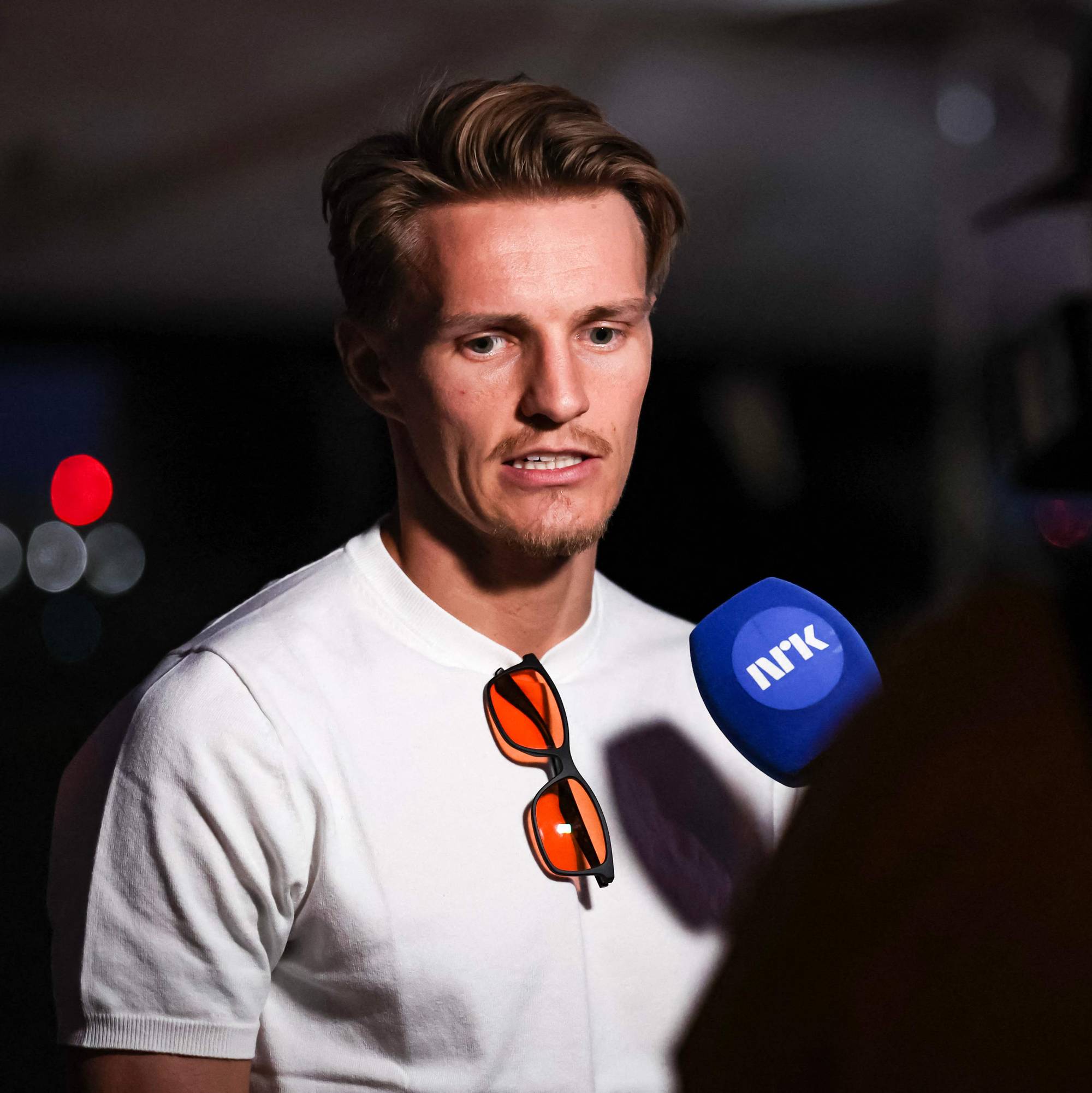 En mann i hvit t-skjorte blir intervjuet av NRK. Han har solbriller hengende på t-skjorten. Bakgrunnen er mørk med utydelige lys. (Bildebeskrivelsen er laget av en KI-tjeneste)