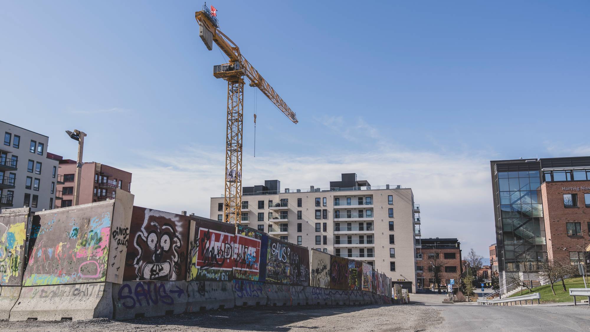 Gul byggekran står ved en byggeplass med graffitimalte betongelementer og boligblokker i bakgrunnen. (Bildebeskrivelsen er laget av en KI-tjeneste)