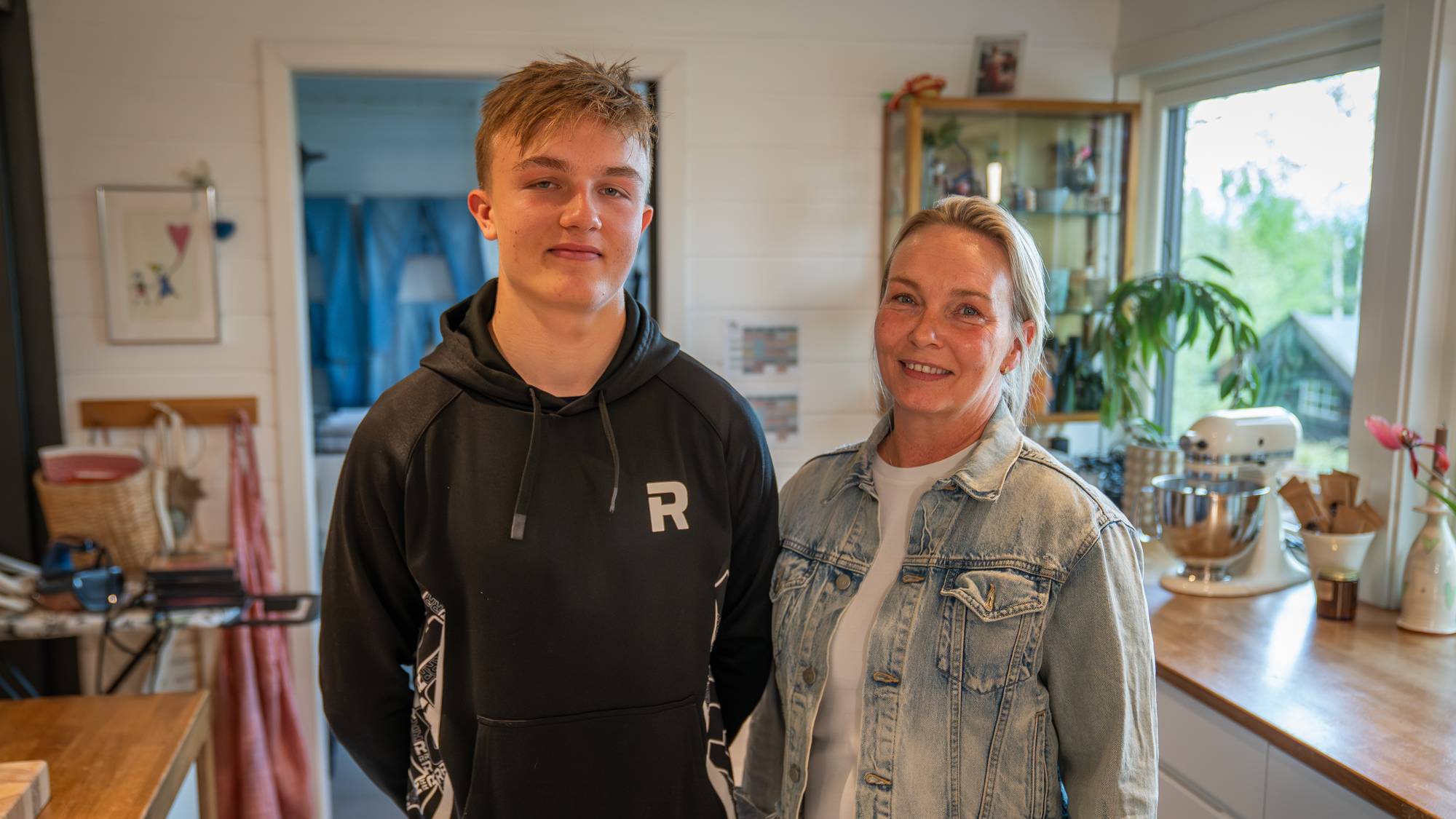 En ung mann og en voksen kvinne står på et kjøkken og smiler. Mannen har på seg en svart hettegenser, og kvinnen har på seg en dongerijakke over en hvit t-skjorte. I bakgrunnen sees kjøkkenutstyr og en døråpning til et annet rom. Bildet er tatt innendørs i et hjem. (Bildebeskrivelsen er laget av en KI-tjeneste)